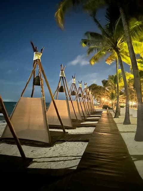 A boardwalk with palm trees and tiki huts outside on a beach.