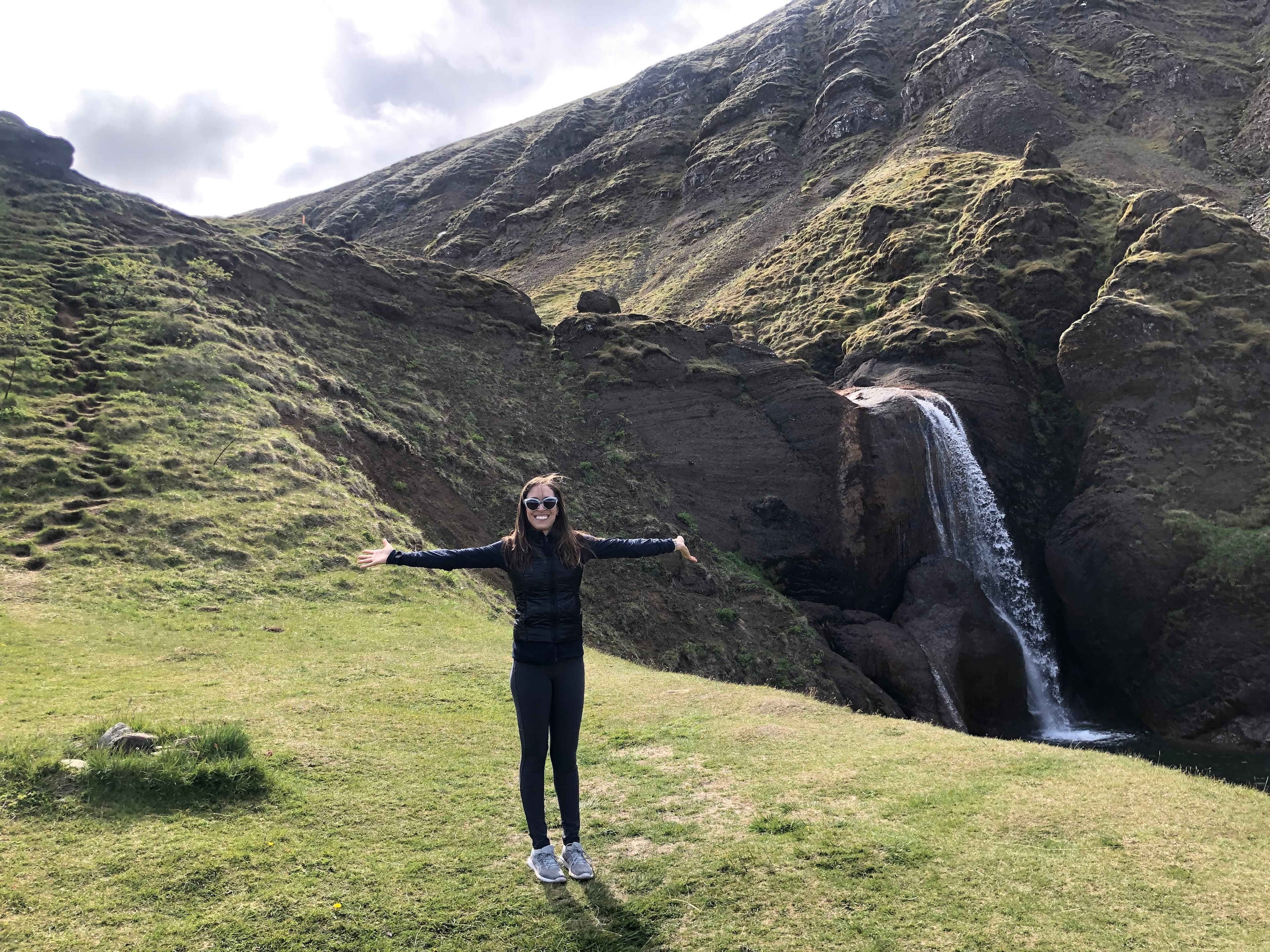 A woman posing outside with her arms outstretched wide in front of a green cliff and waterfall.