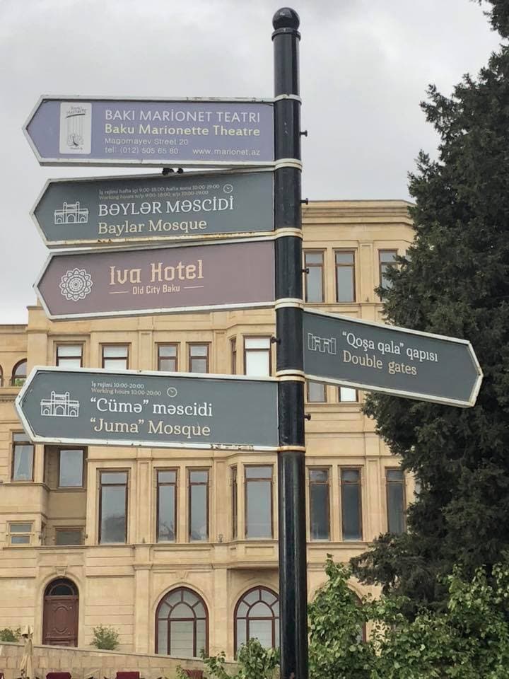 A view of signs during a sunny day with foliage and buildings in the distance.