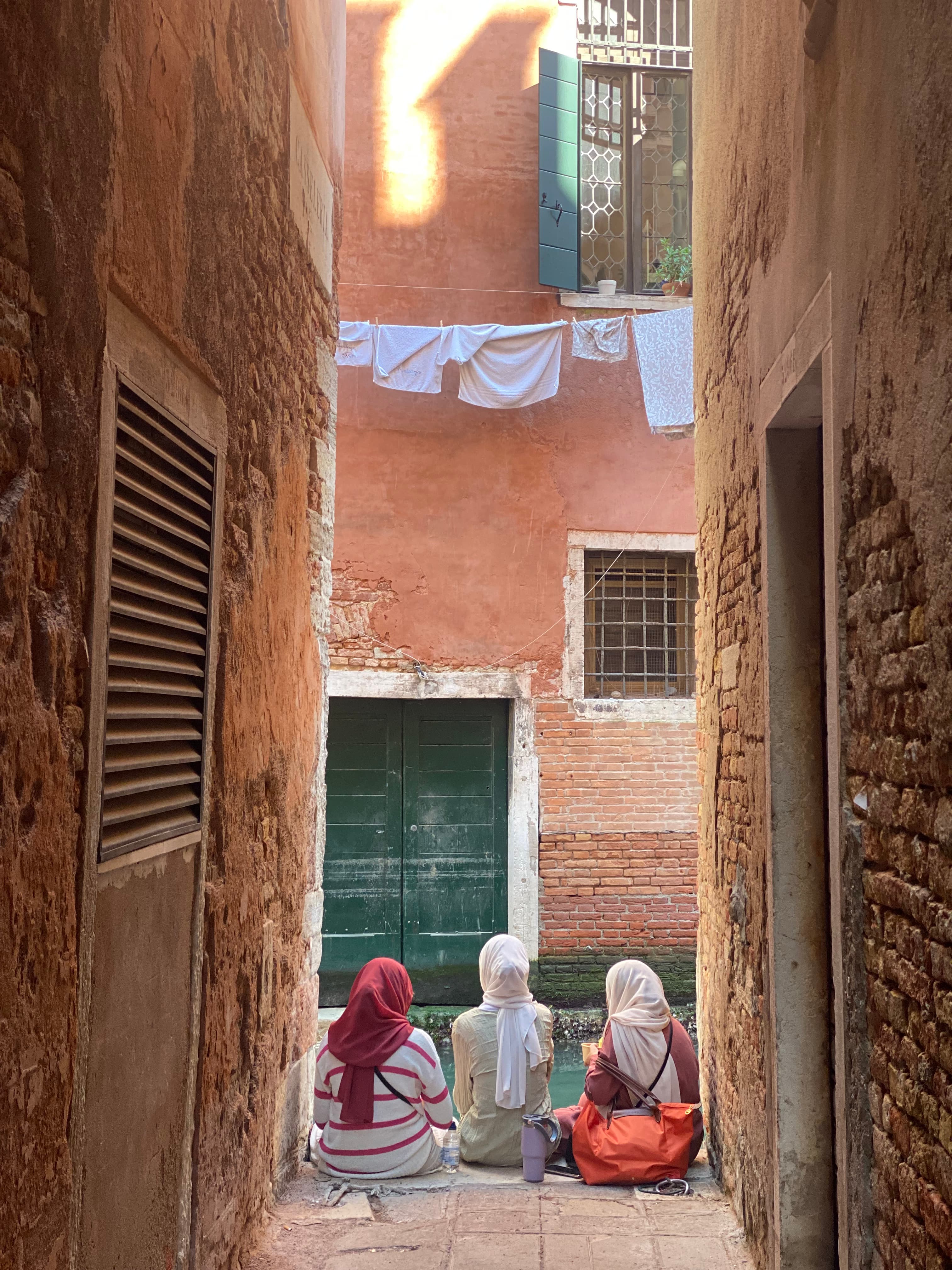 Three people with headscarves sit in a narrow walkway with windows and laundry hanging above them.