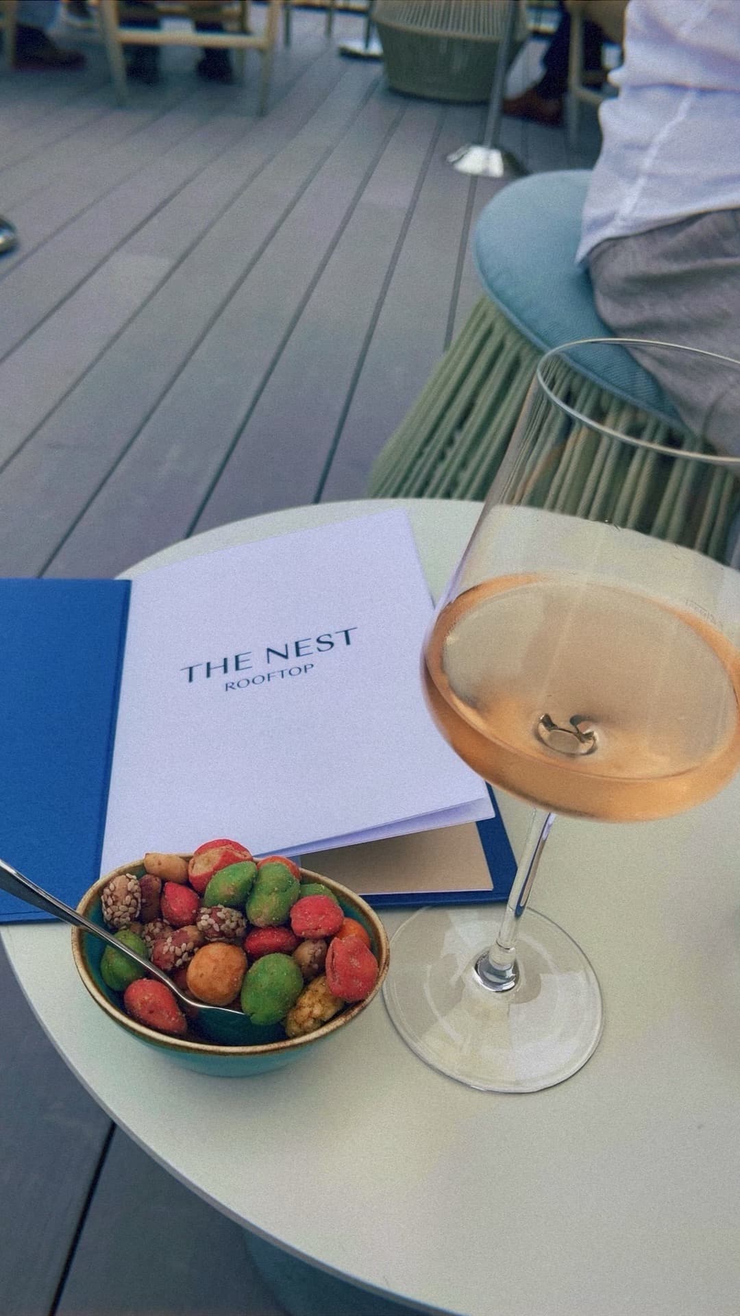 Glass of rose and small bowl of snacks beside a blue menu on a white circular table
