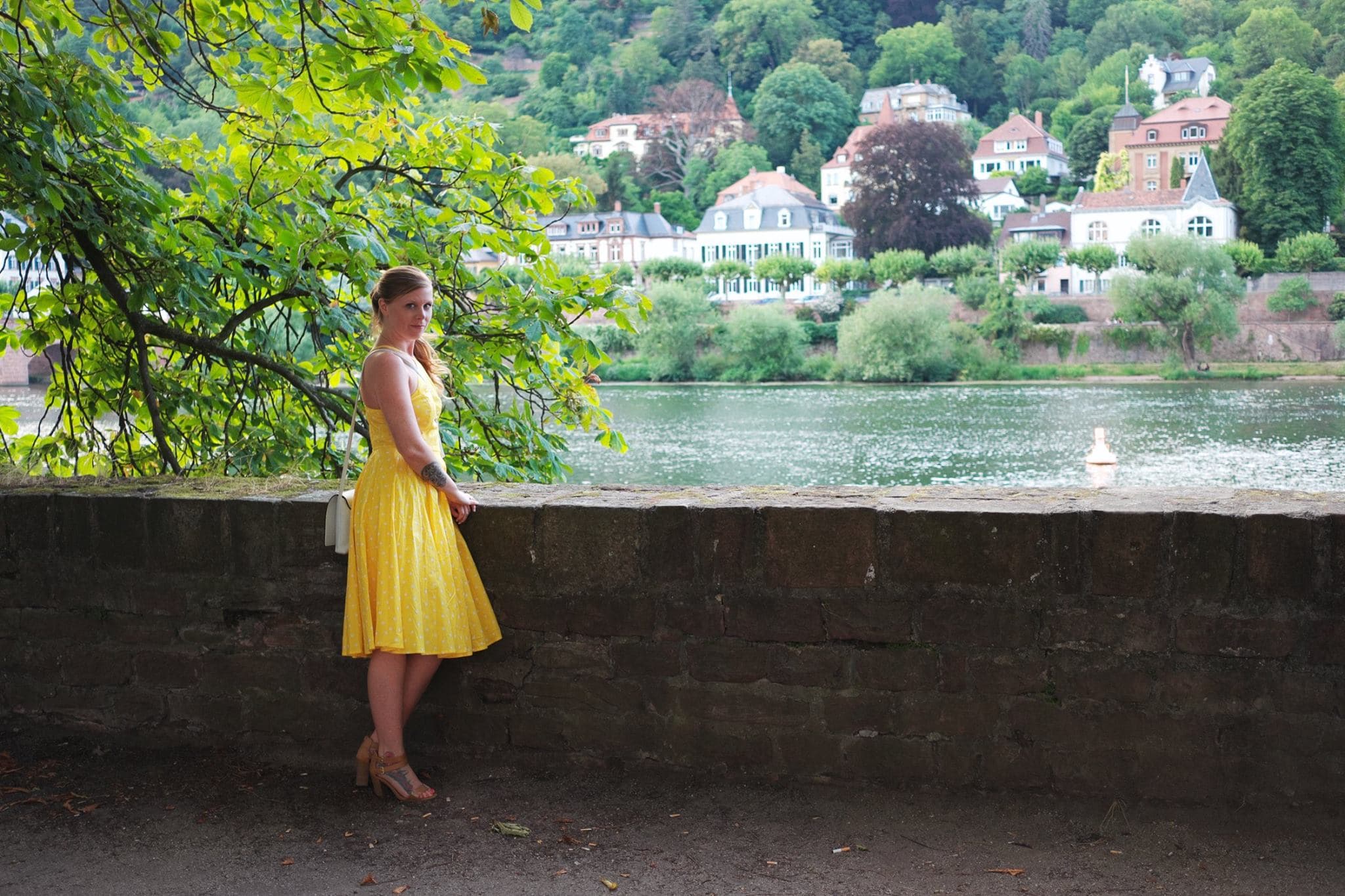 Jeanette posing against a brick ledge wearing a yellow dress with the city and water in the background.