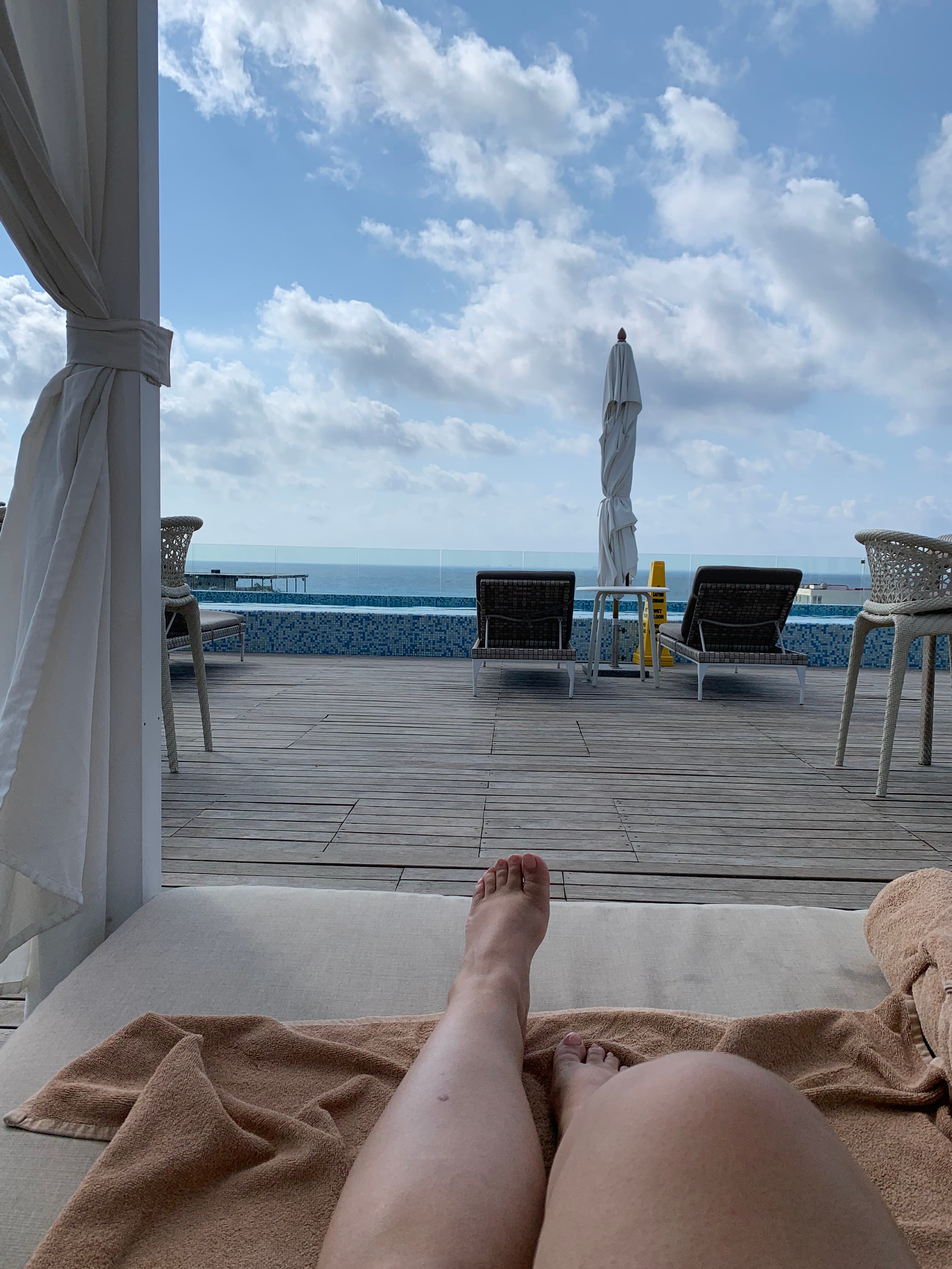 A person's legs laying on a sun bed next to a pool during the daytime