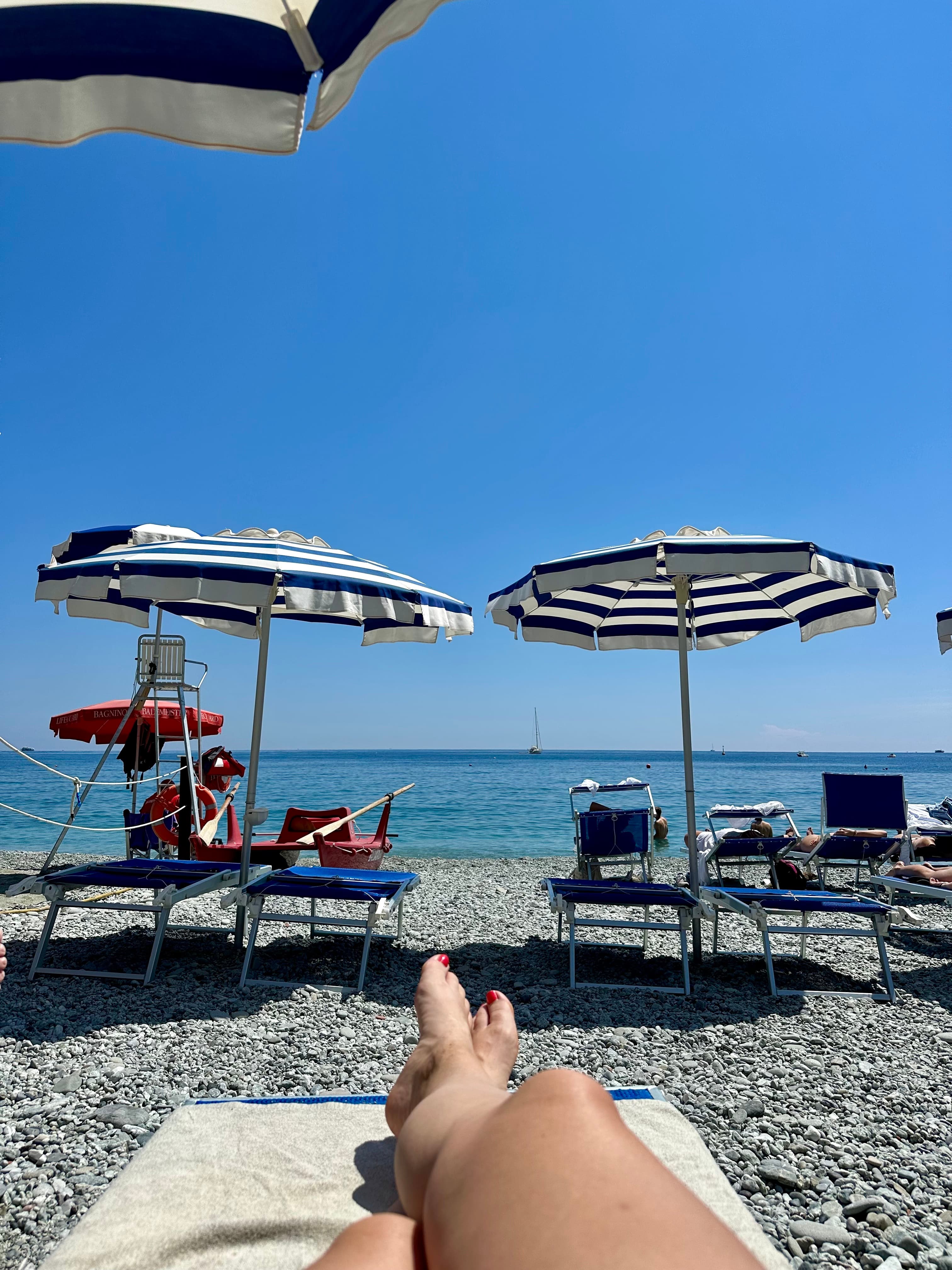 A view of a person's legs on the beach, relaxed under striped umbrellas, surrounded by sand and gentle waves.