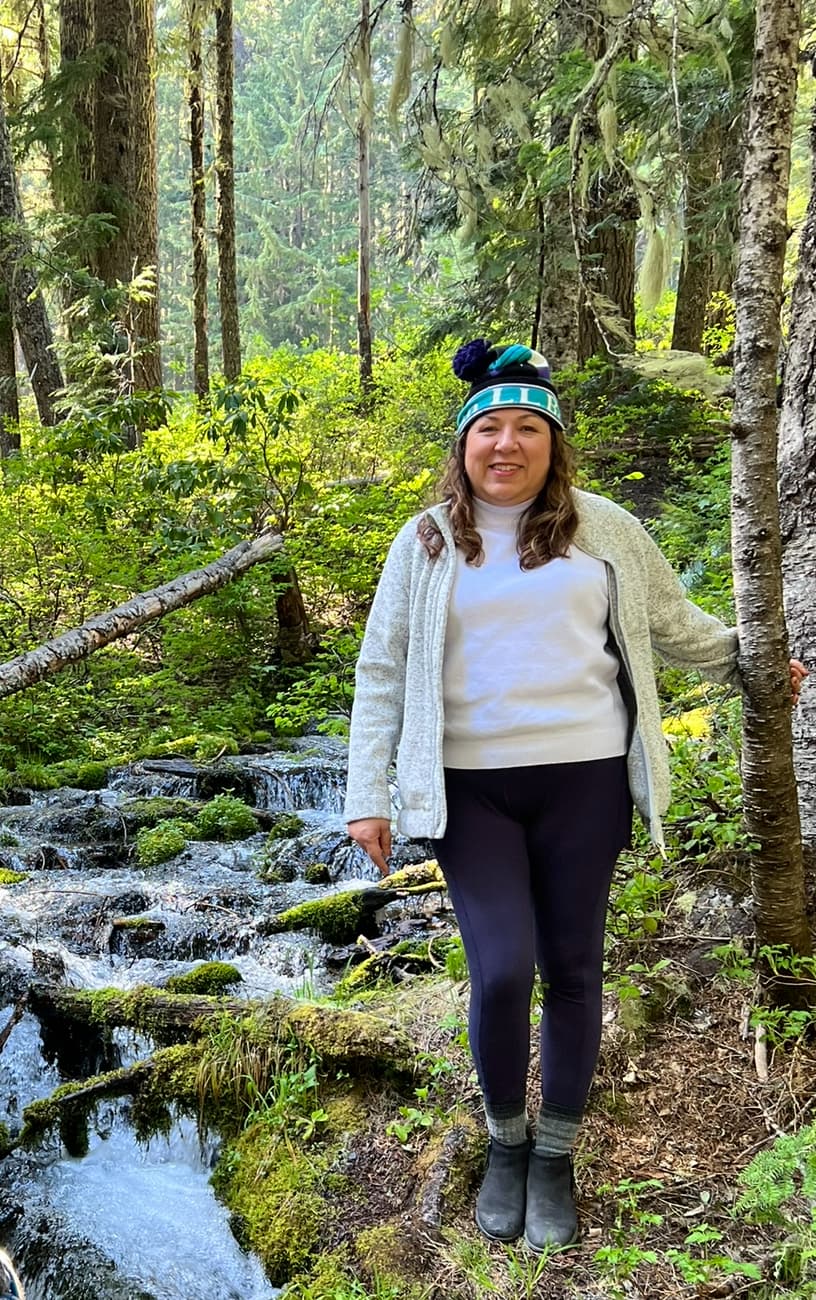 A woman wearing a sweater and a blue hat stands beside a serene stream, enjoying the tranquil natural surroundings.