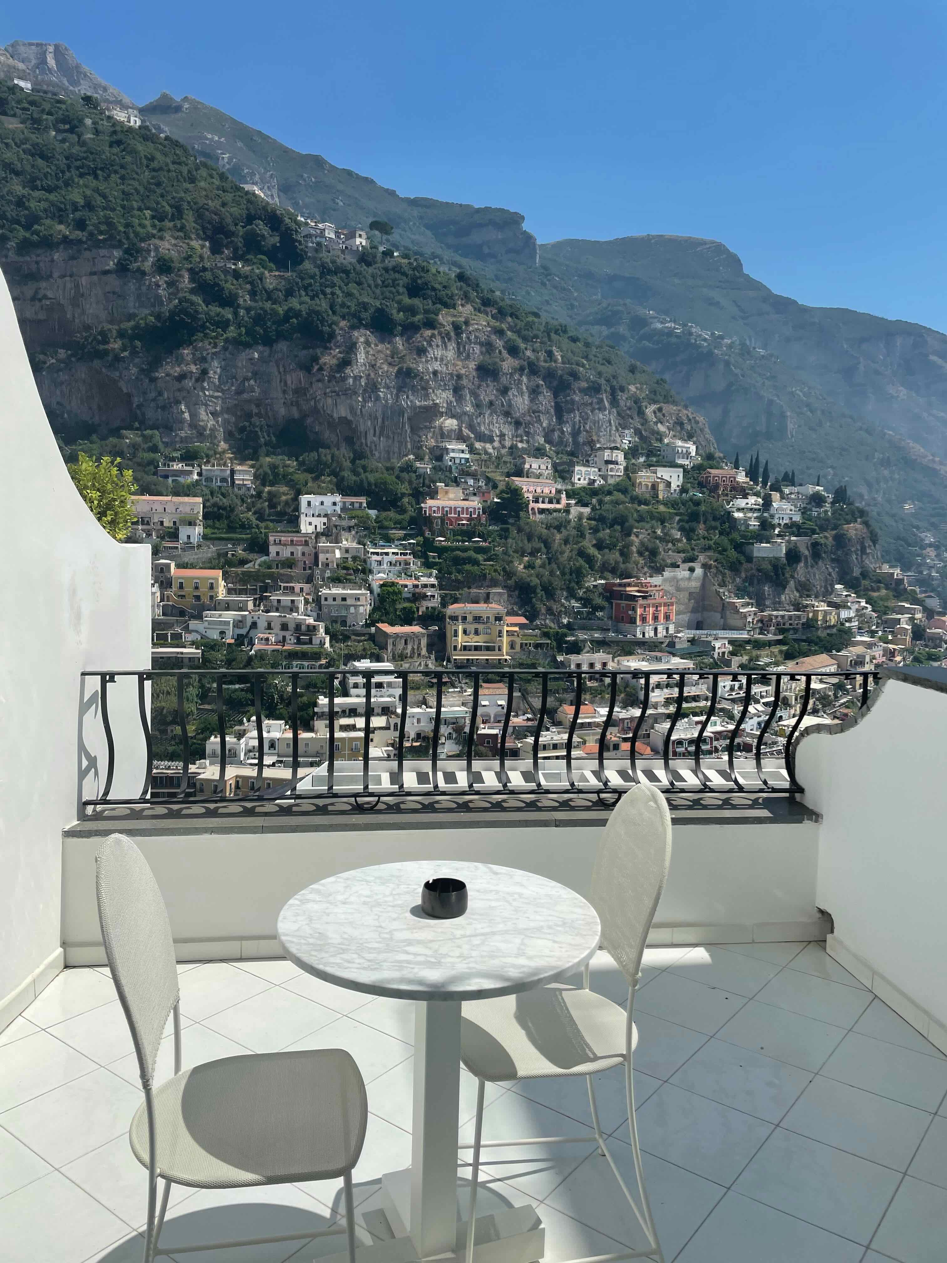 View of two white chairs and a circular table on a balcony overlooking a beautiful cliffside town