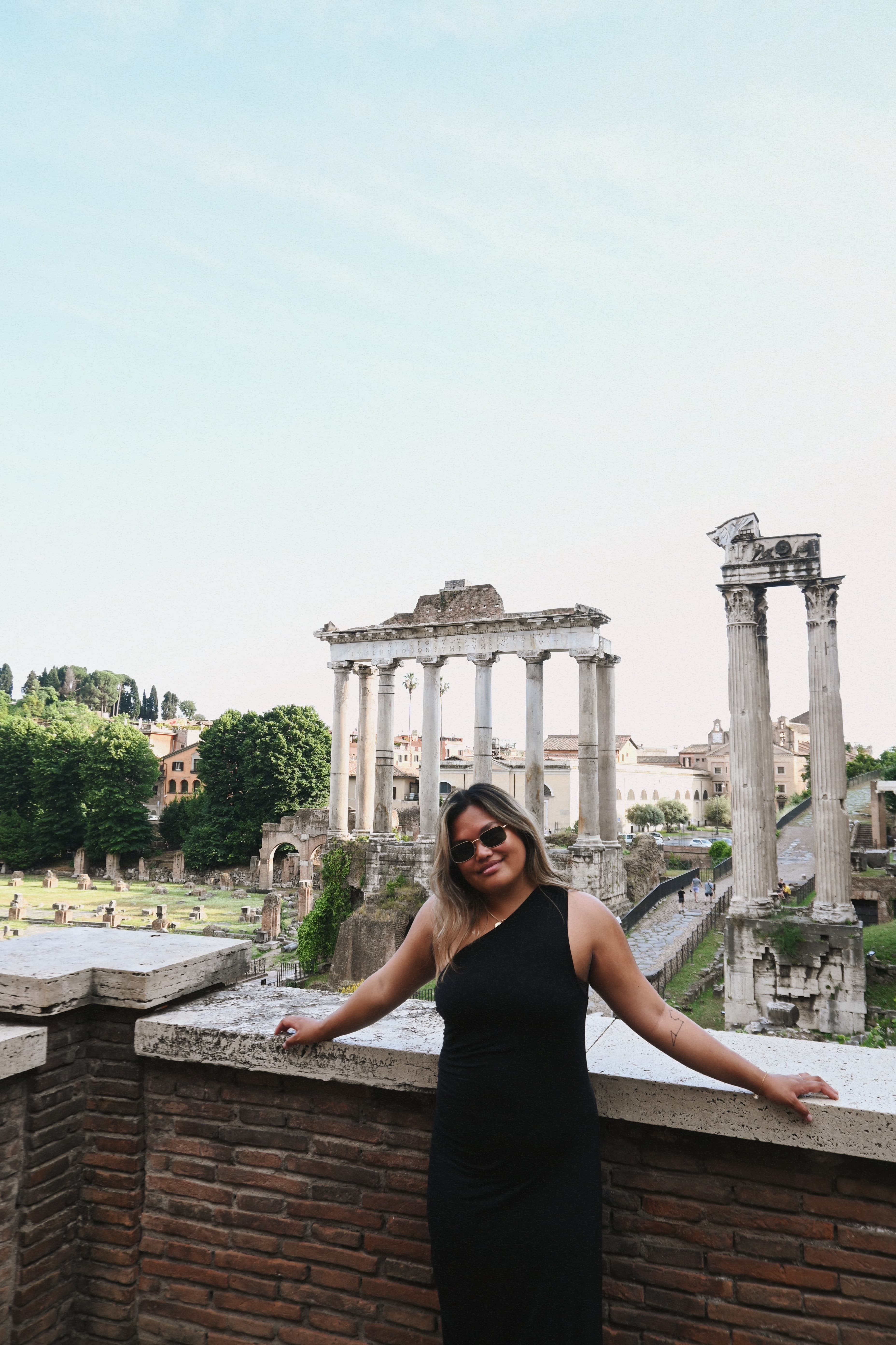 Kalei wearing a black dress poses on a wall, with the Roman Forum's ancient ruins visible in the background.