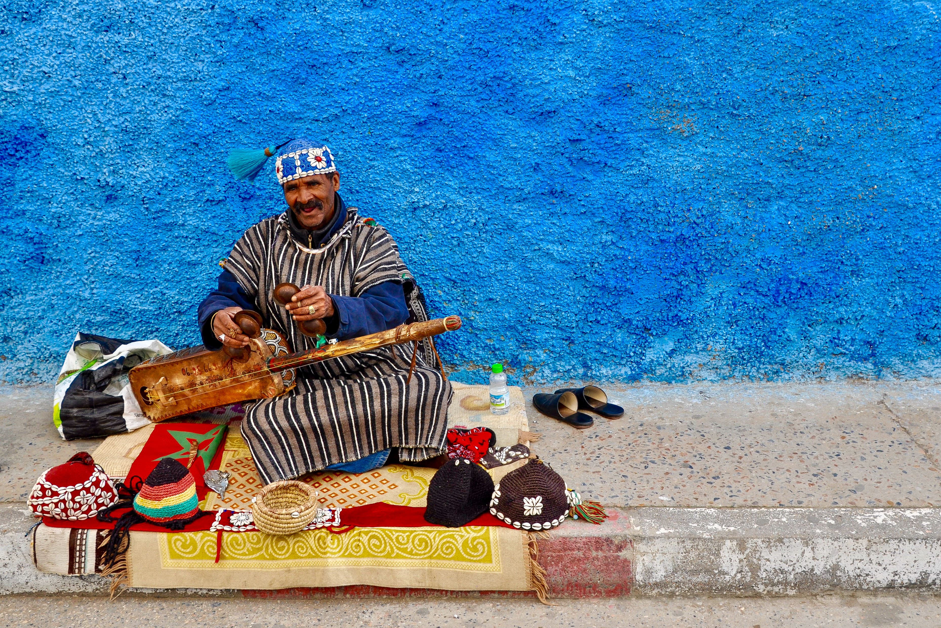 A man sitting along a sidewalk with a bright blue backdrop with items surrounding him.