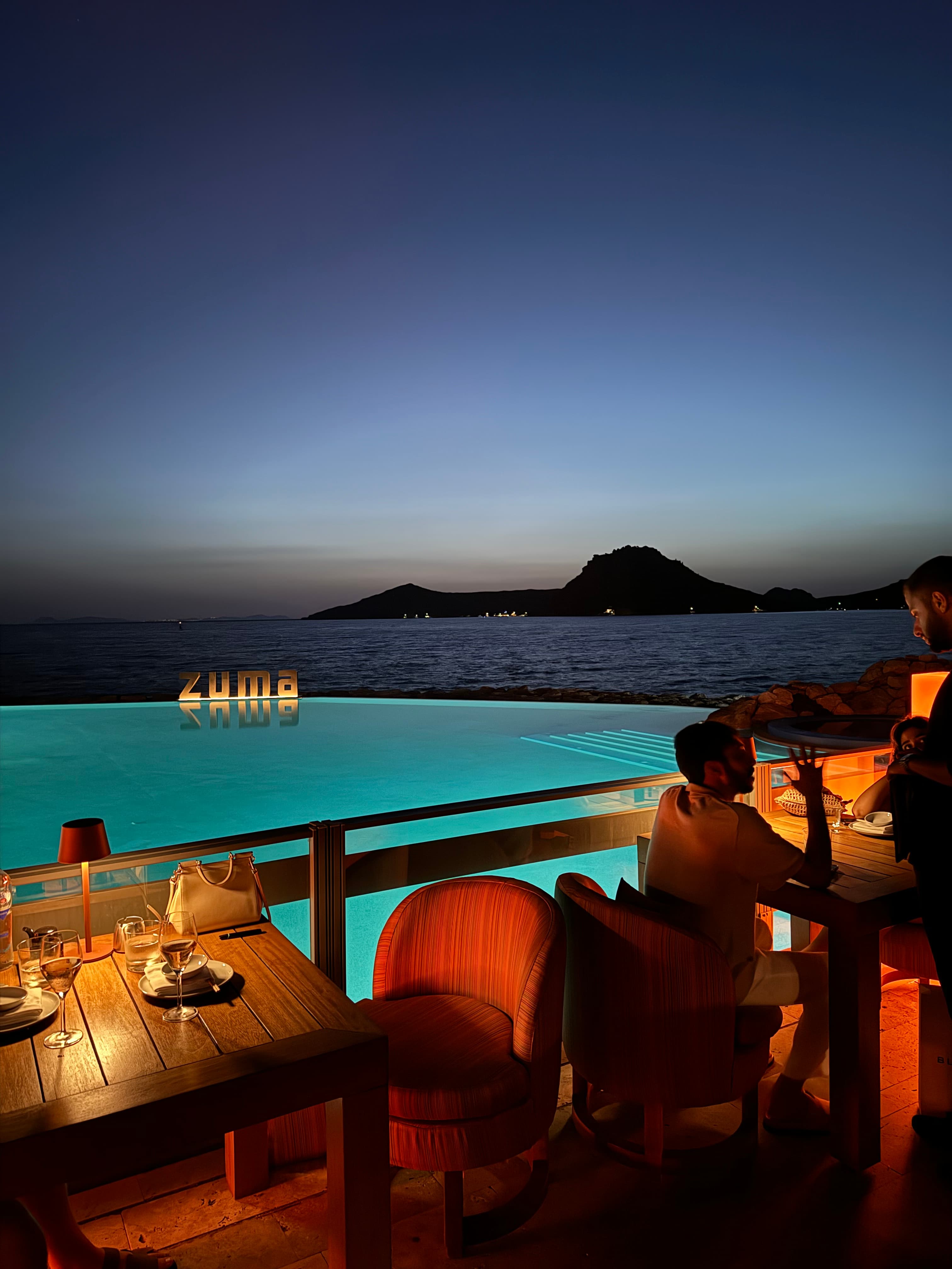 View of people dining in dim light by a resort swimming pool overlooking the sea at dusk