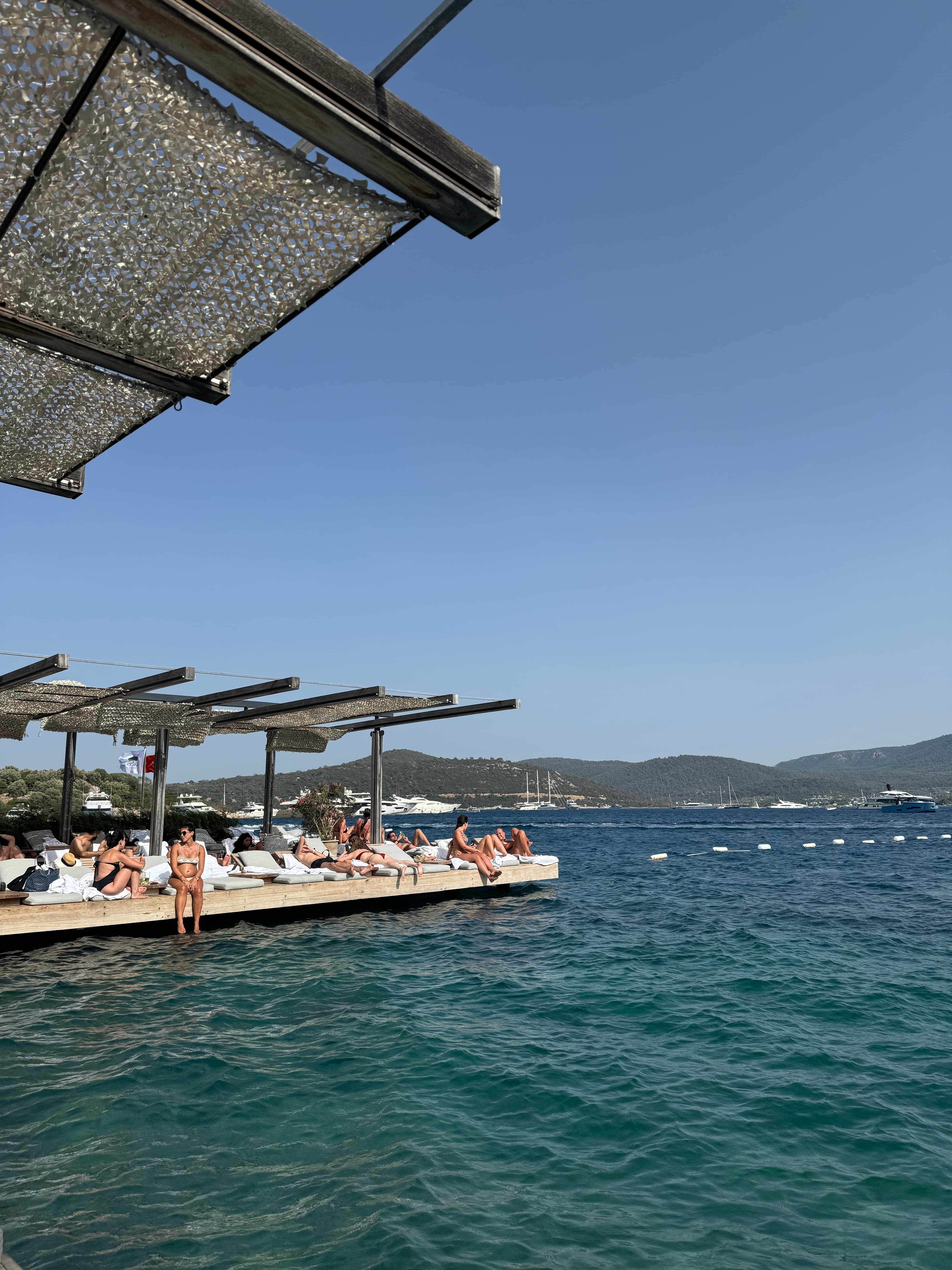 View of sunbathers on lounges on a platform by the sea under clear skies