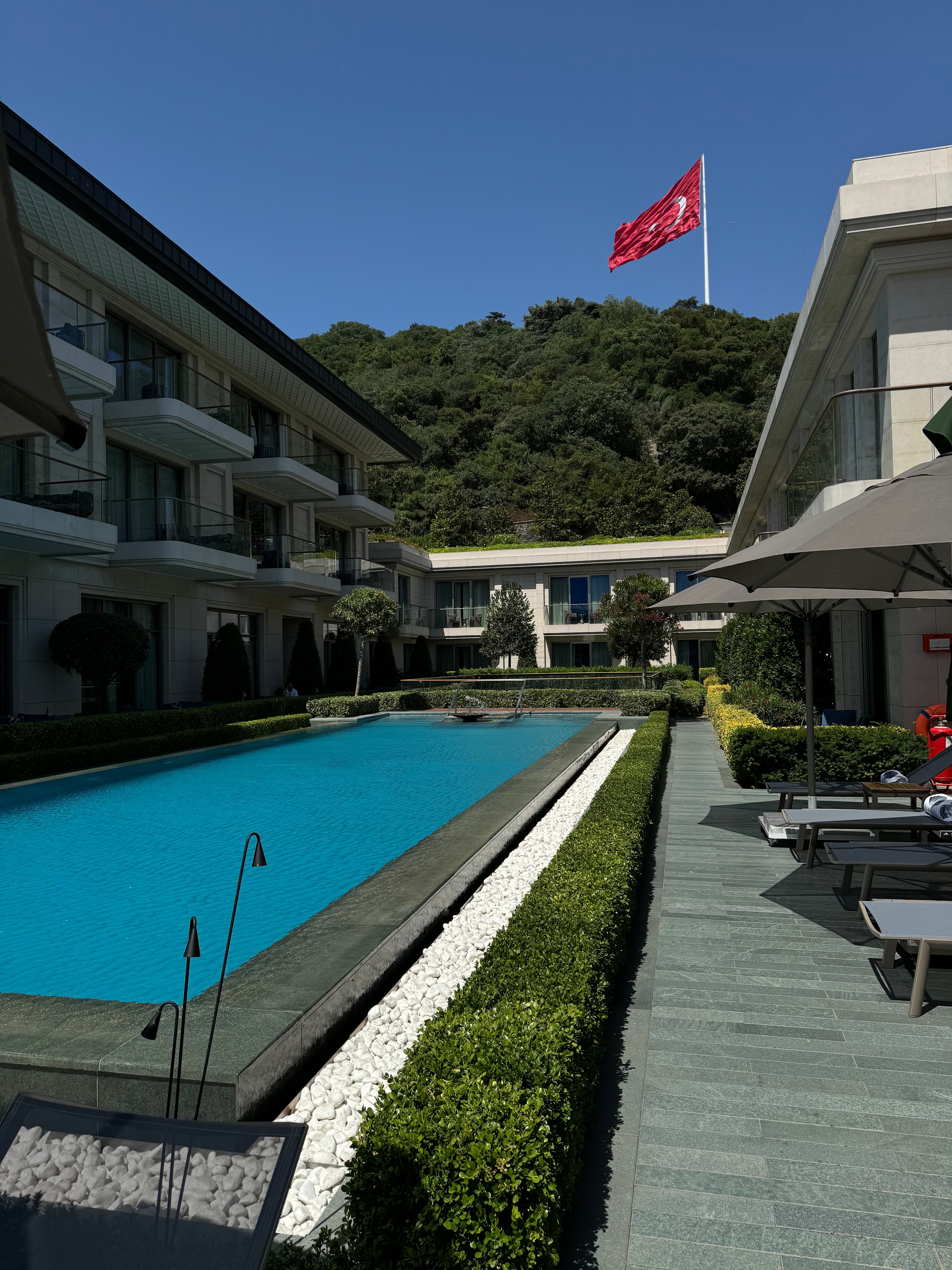 View of a long rectangular swimming pool at a hotel with a red flag flying above the treetops