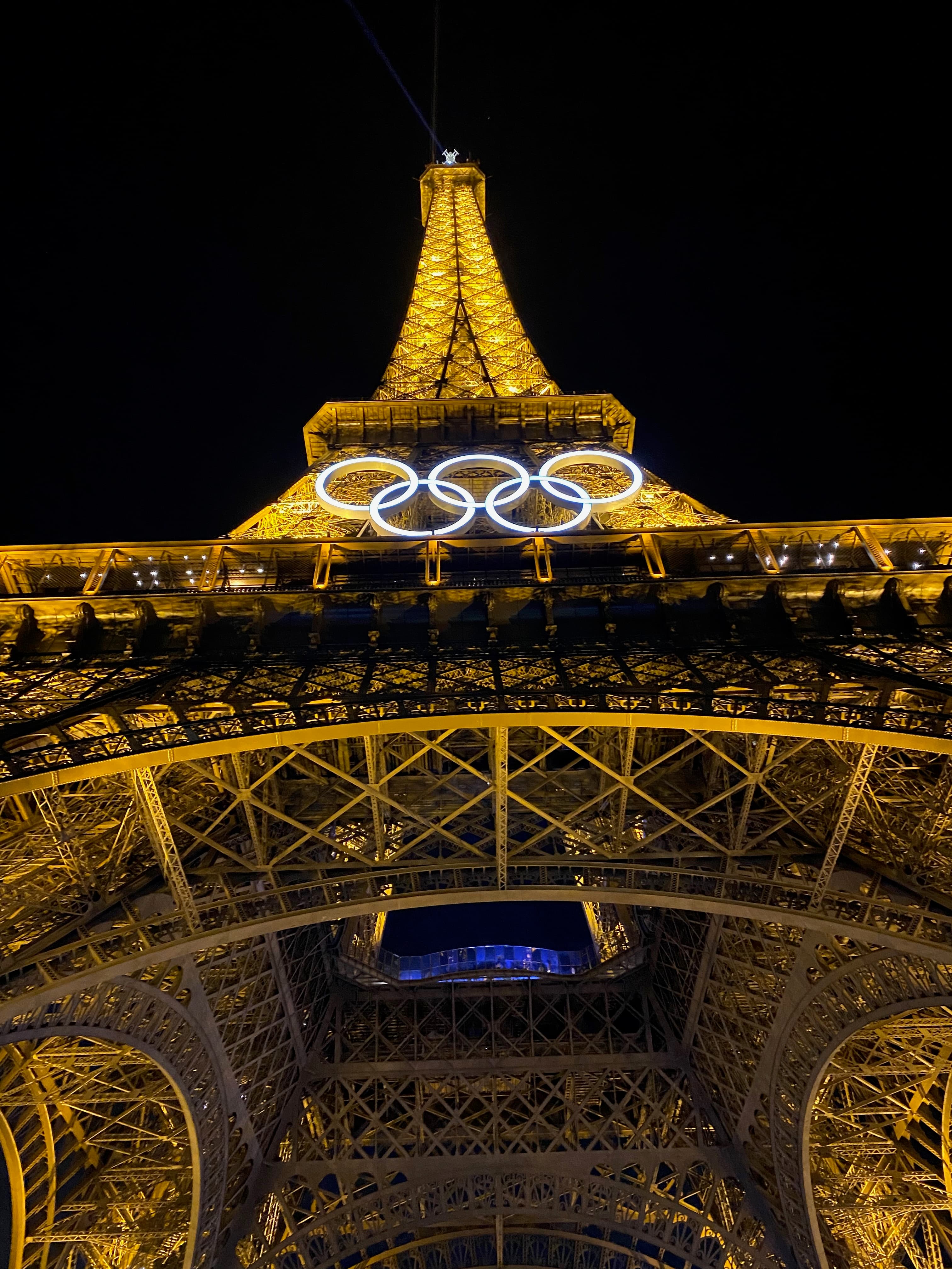 View from the bottom of the Eiffel Tower of the structure lit up at night with the Olympic rings hanging in front