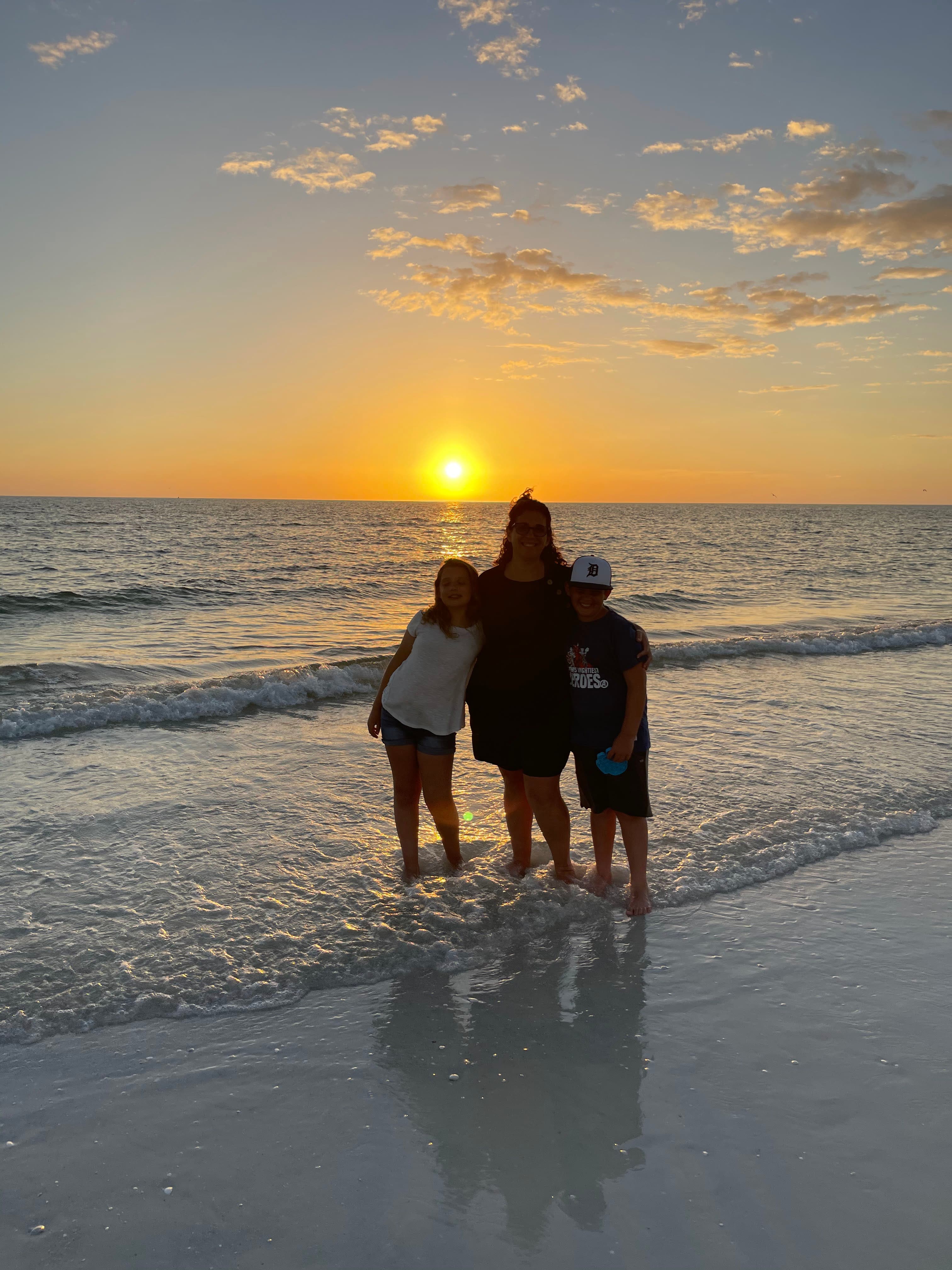 An image of a person standing on the beach with two children on each side of them in front of a sunset and ocean.