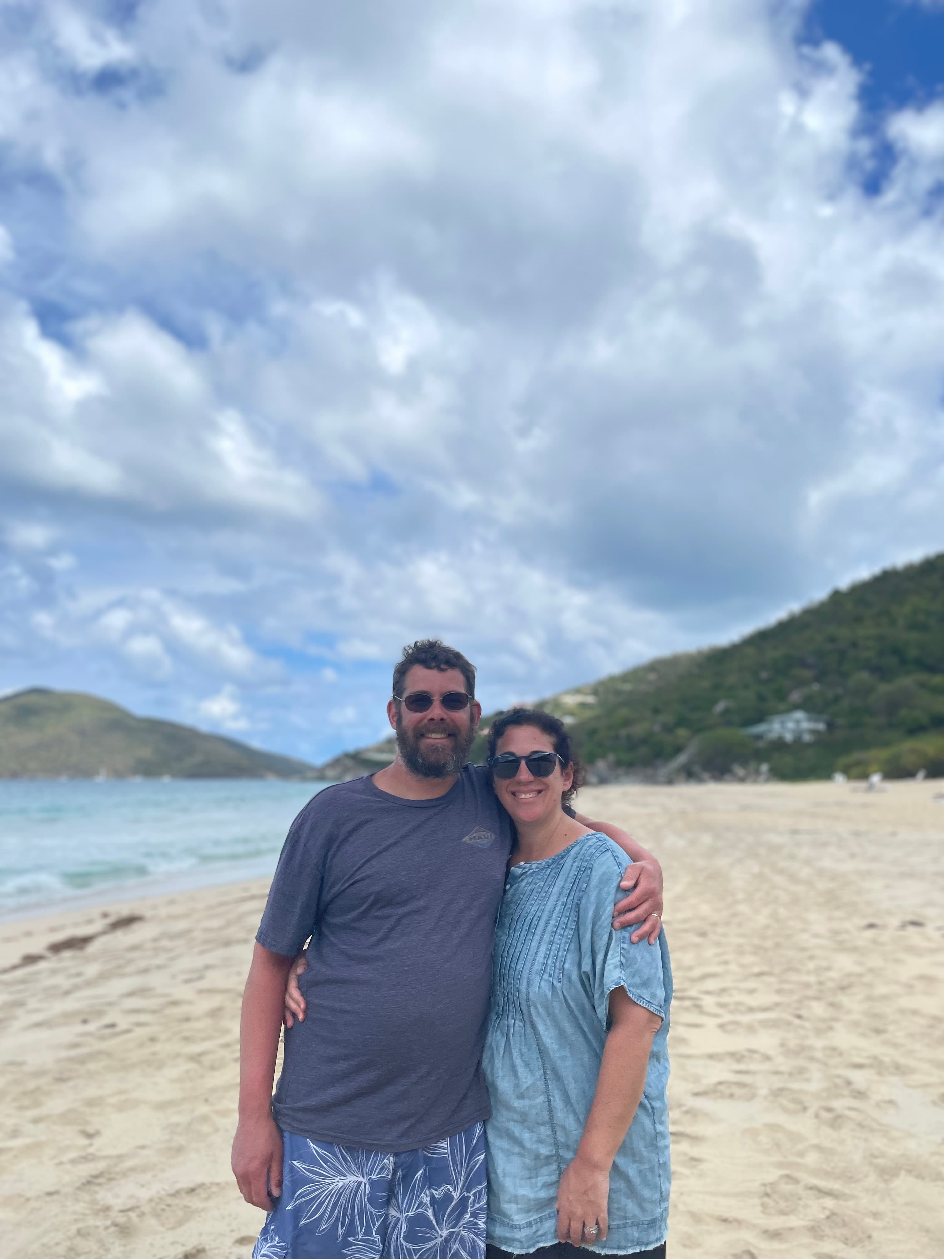 A couple posing for a photo on the beach with island-like terrain in the background. They are both wearing cool-toned clothing and black sunglasses.