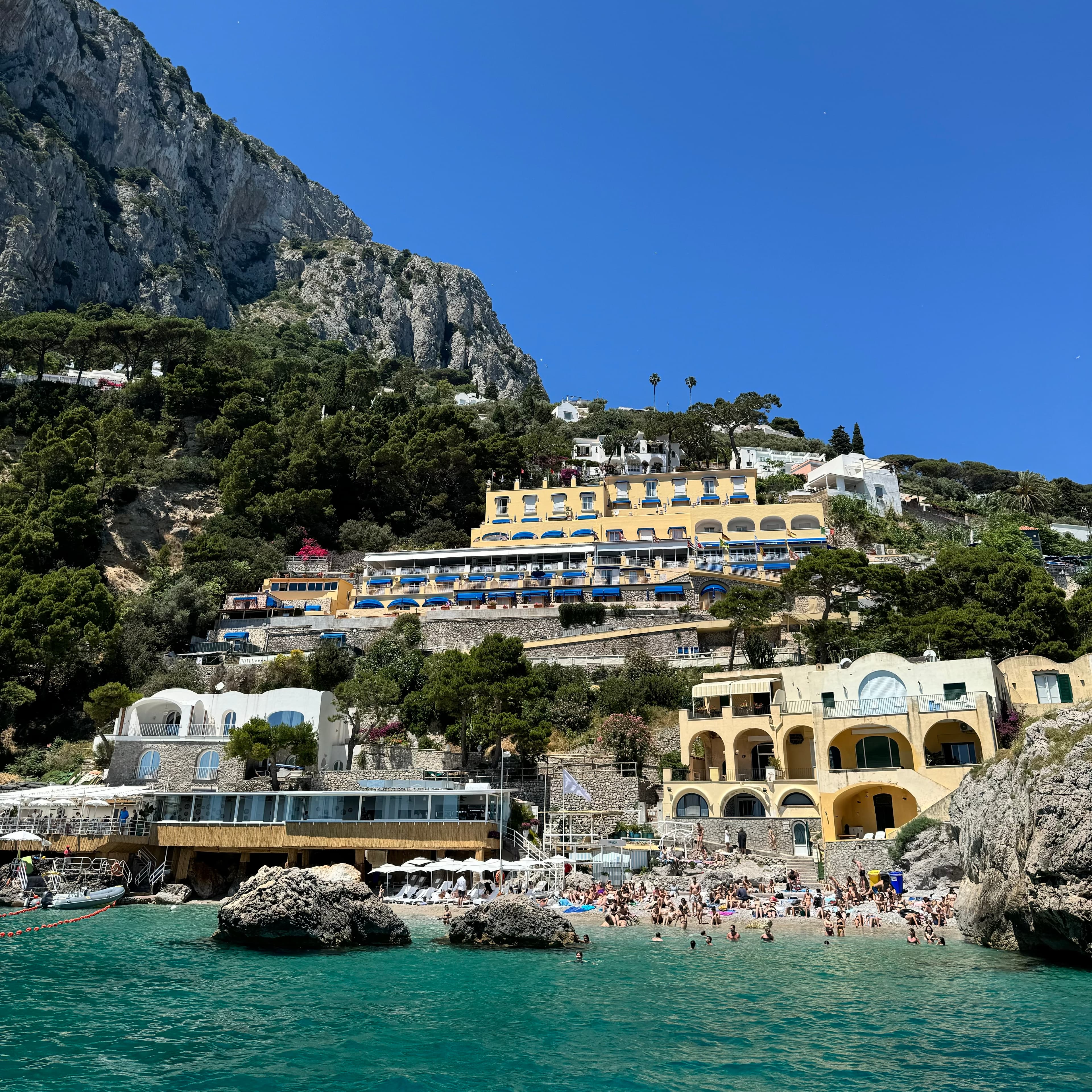 View from the sea of a small rocky beach crowded with sunbathers and buildings on the cliffside above