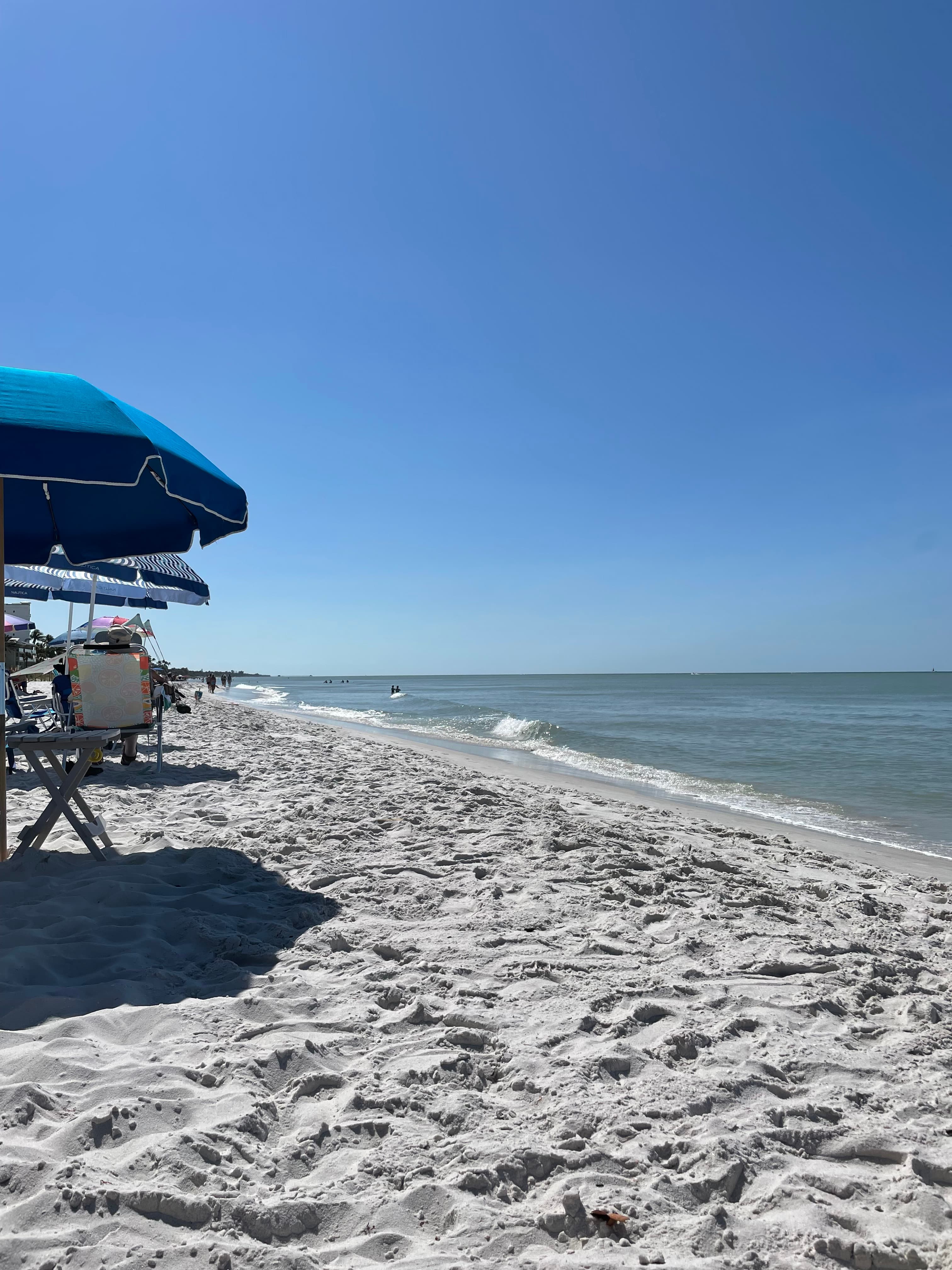 A view of a white sand beach, with lounge chairs and umbrellas and calm waves washing on the shore on a clear blue day.