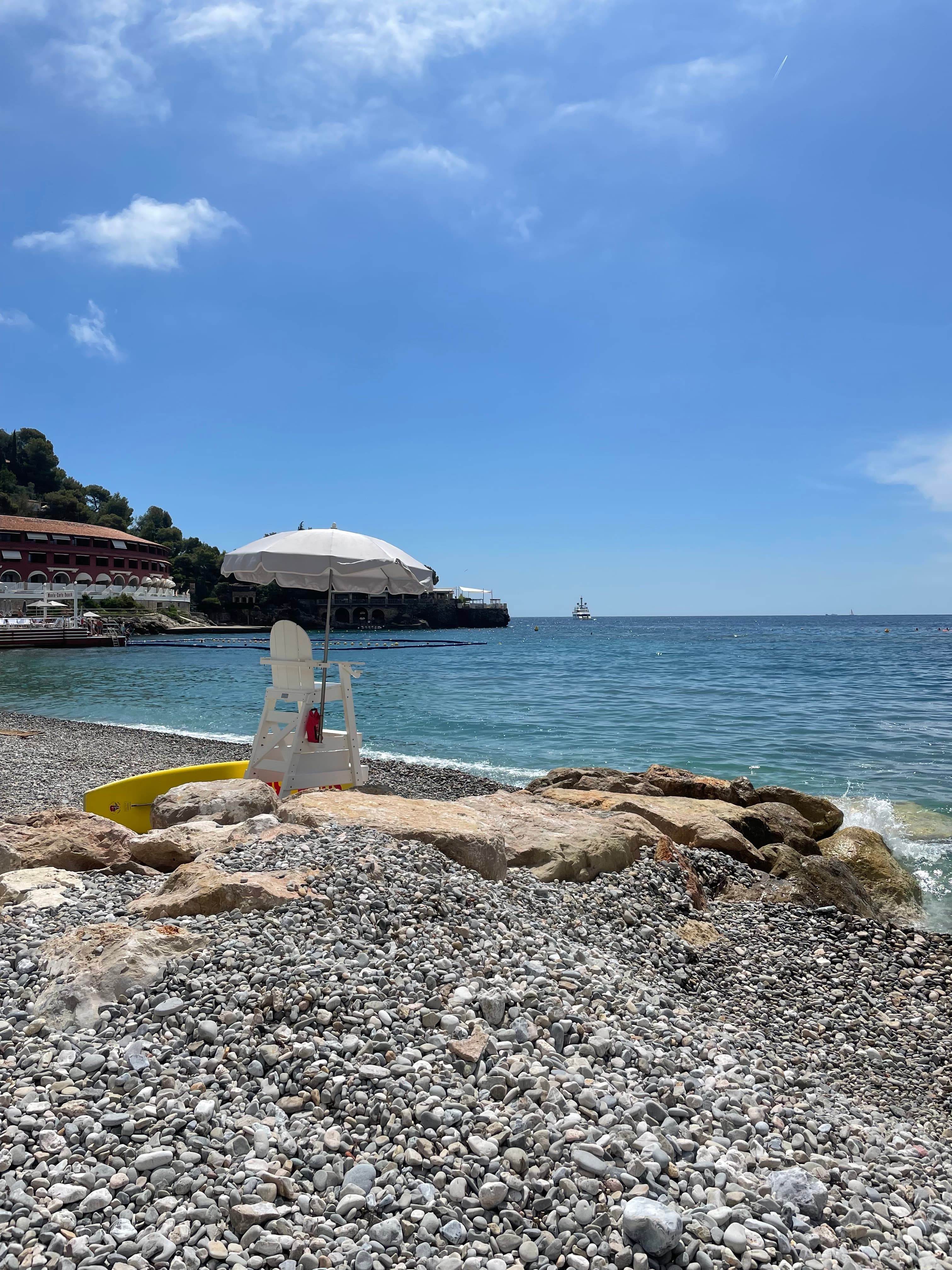 A view of the lifeguard stand covered by an umbrella with a yellow surfboard leaning against it on a rocky beach during the day.