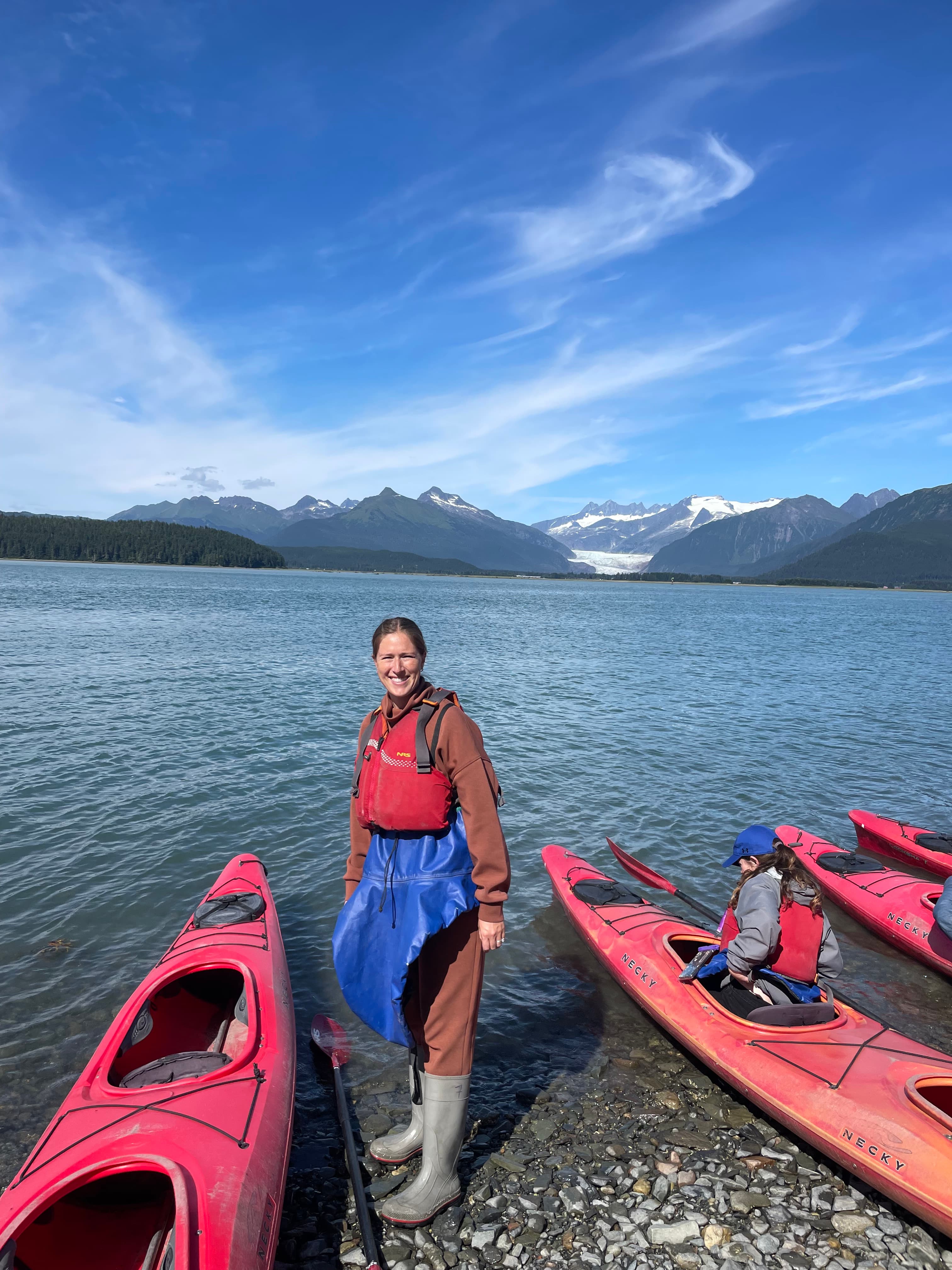 Advisor standing between two red canoes on the bank of a rocky beach and calm water with snowy mountains in the distance during the day.