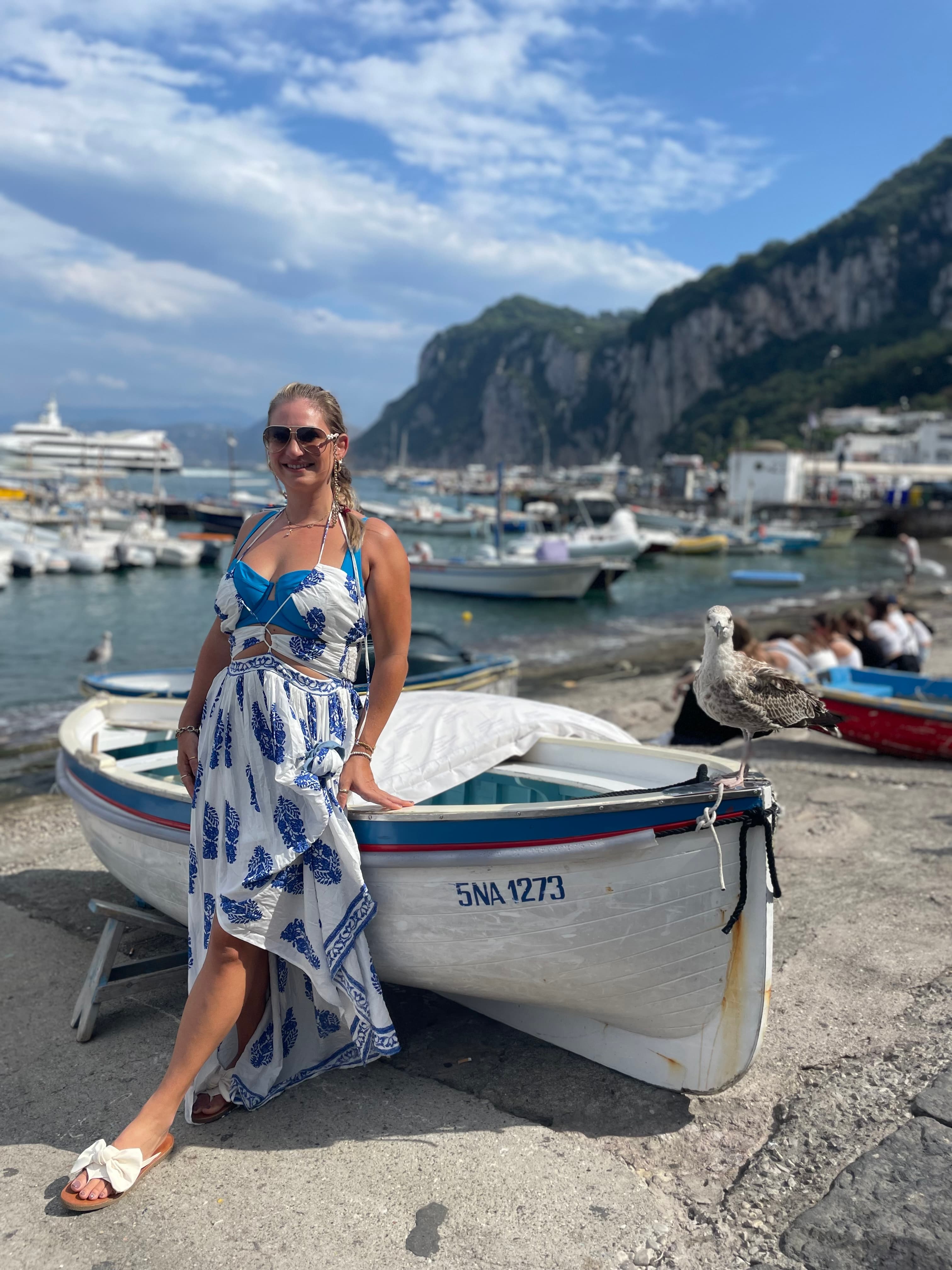 Advisor in a blue and white dress leaning against a small boat on shore in Capri, Italy
