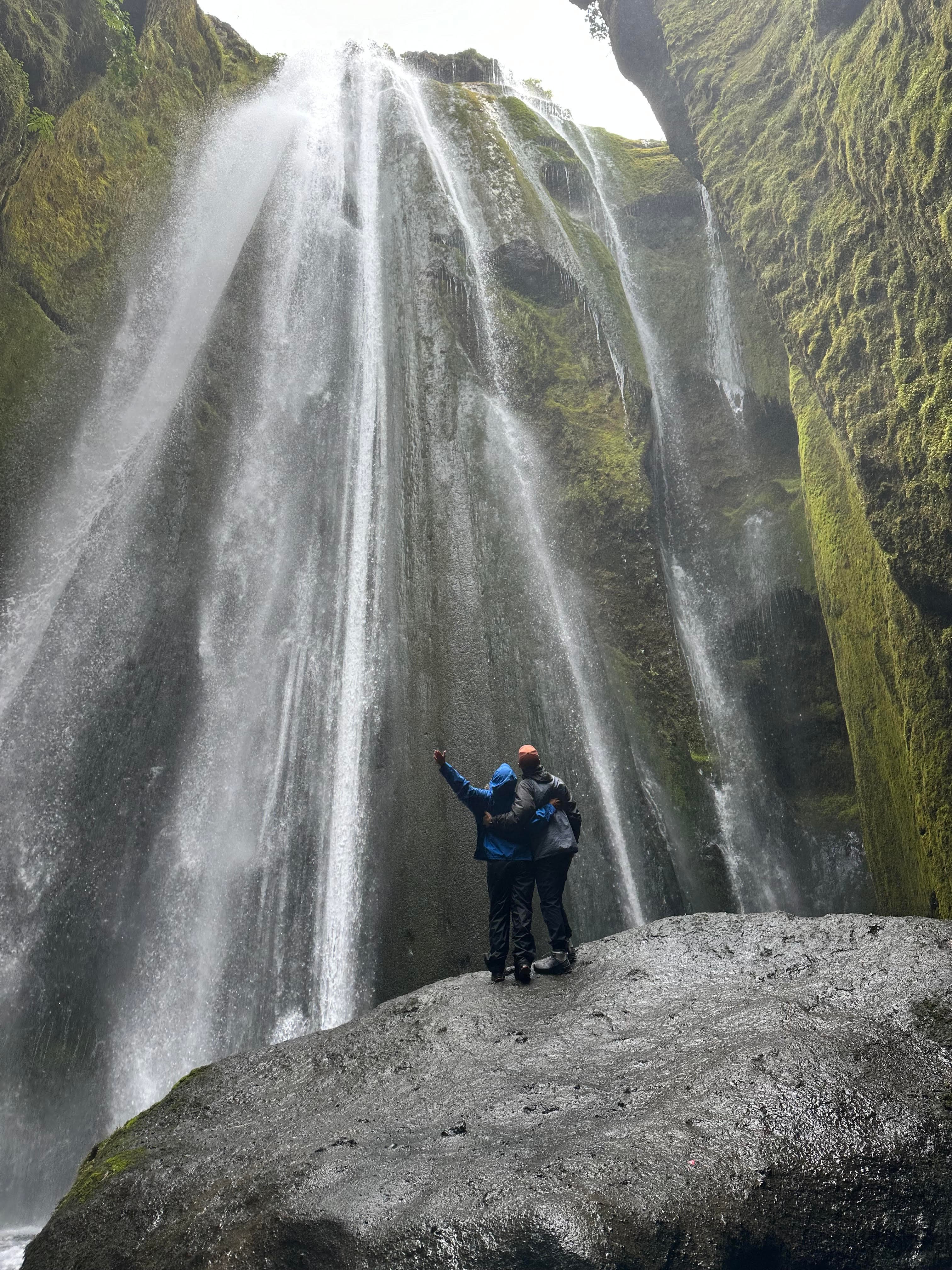 Advisor and another person standing on a large rock at the base of a waterfall on a cloudy day