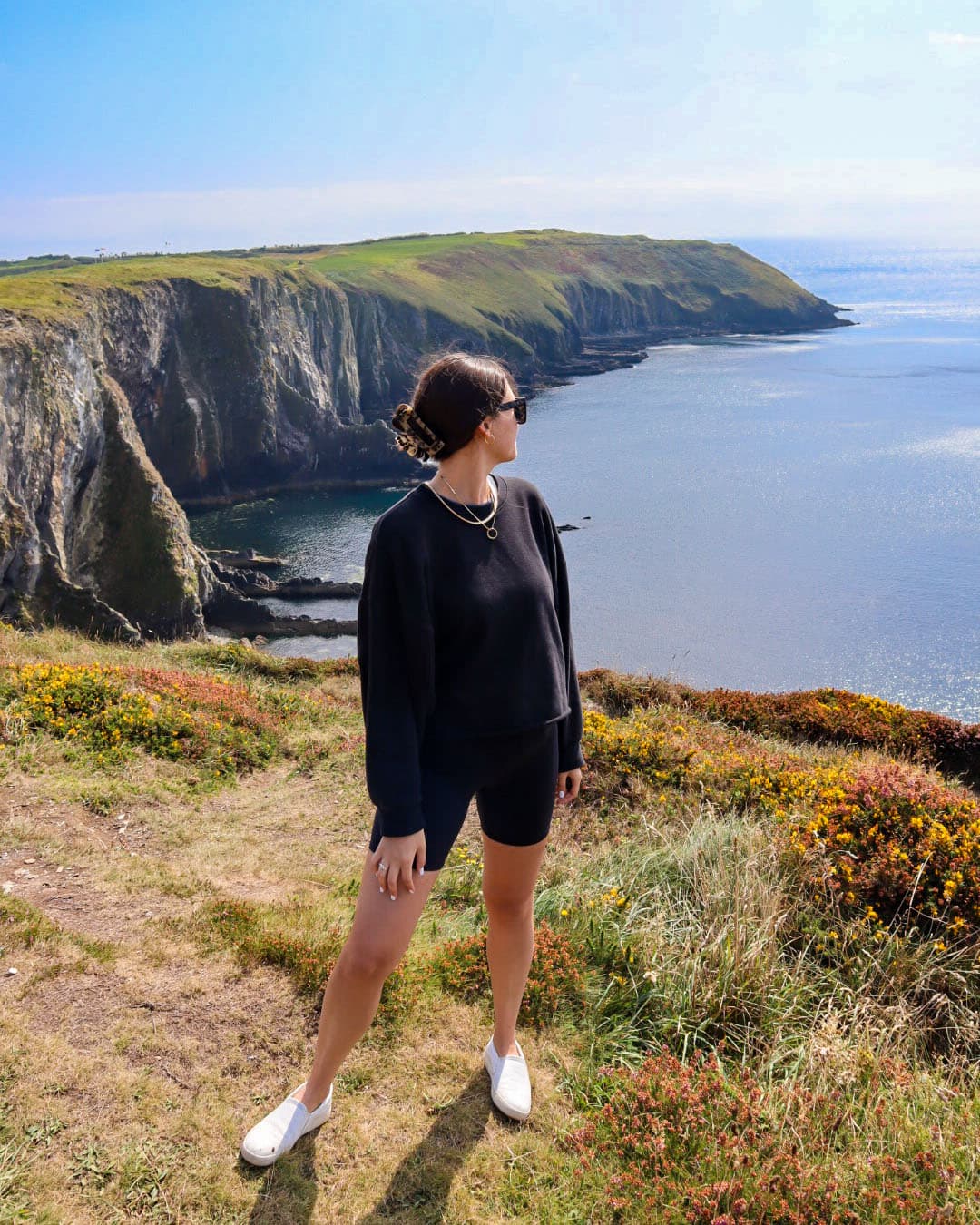 Advisor dressed in all black on a hike with a beautiful coastline of dramatic cliffs behind her