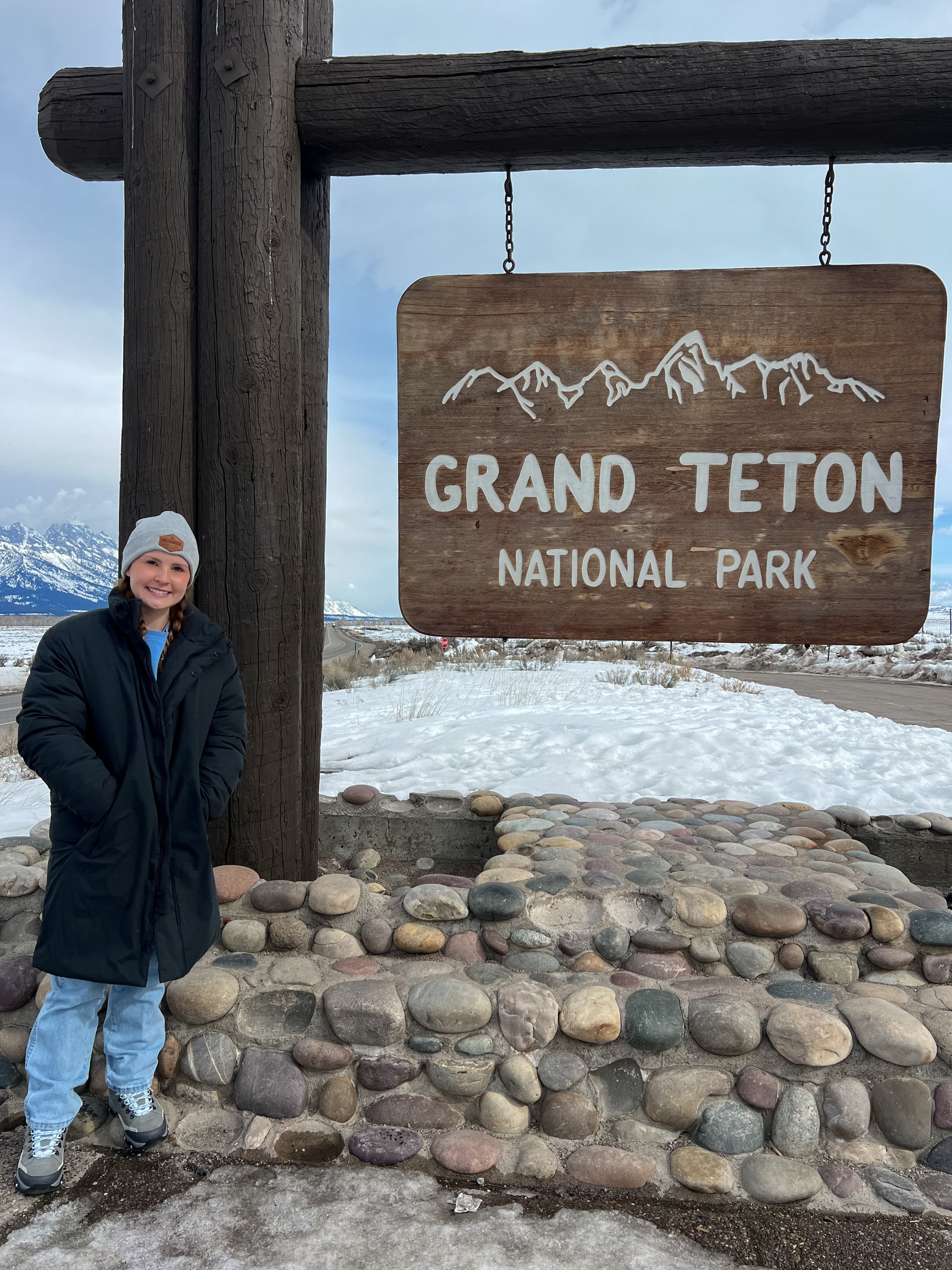Blair posing next to a sign that reads, "Grand Teton National Park", during the daytime.