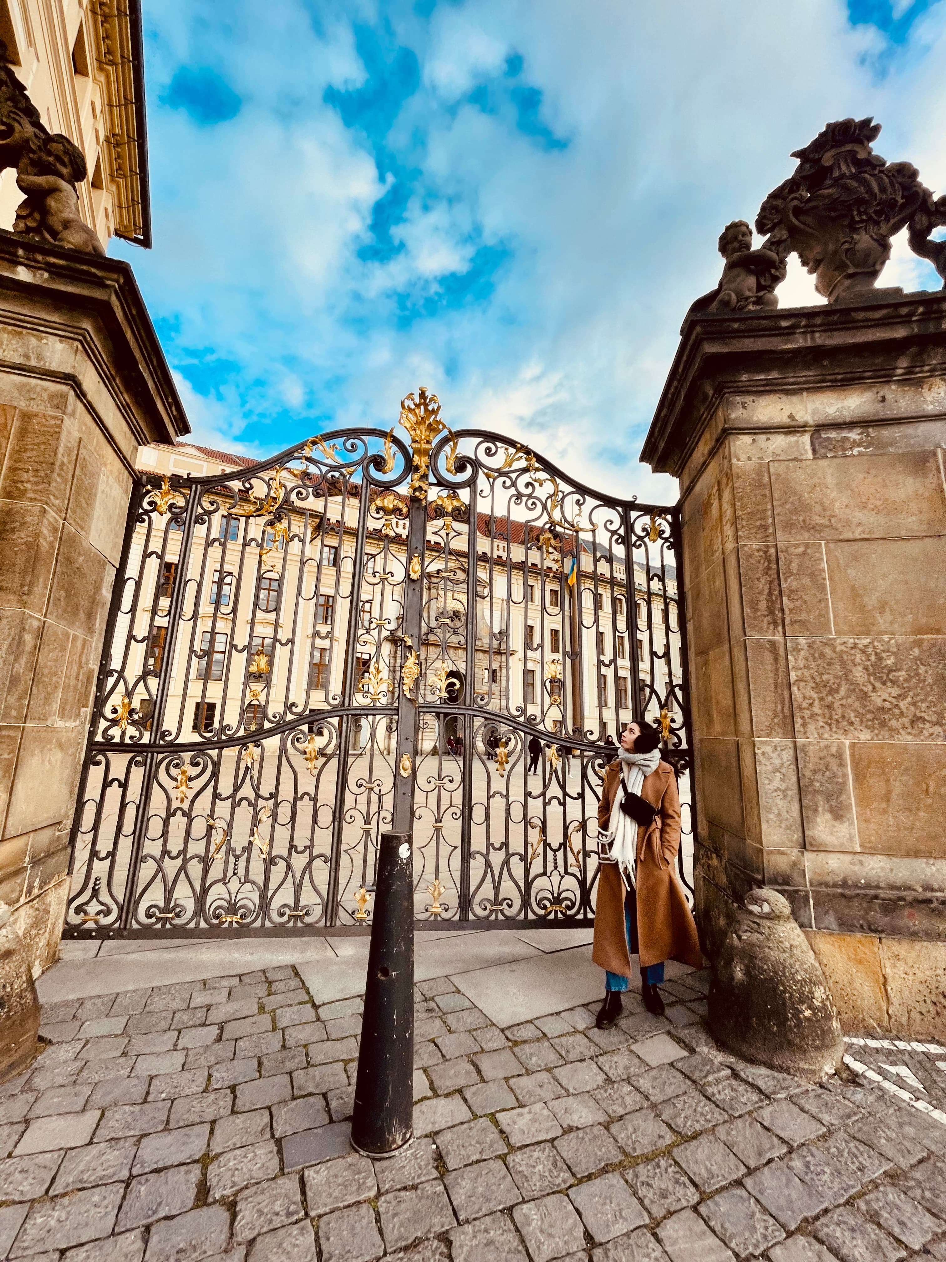 Katie wearing a coat stands poised in front of an ornate gate during the daytime.