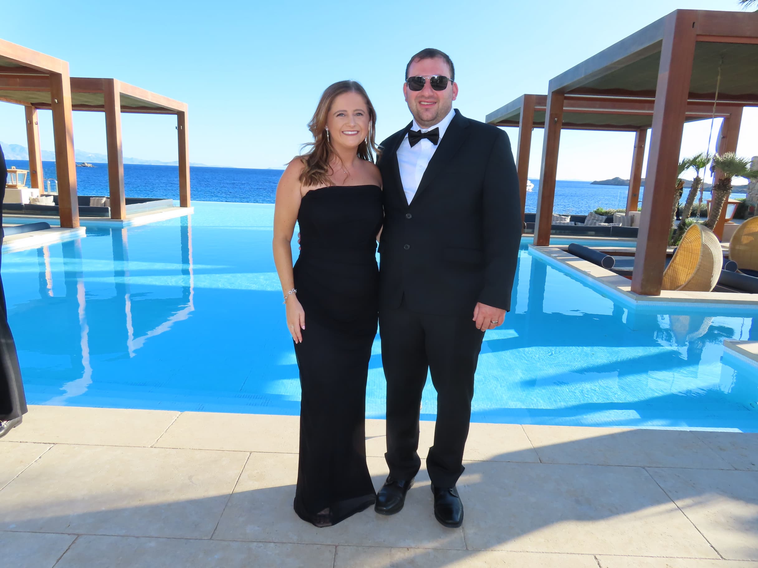 Advisor and husband in black tie dress posing side by side in front of a resort pool on a sunny afternoon