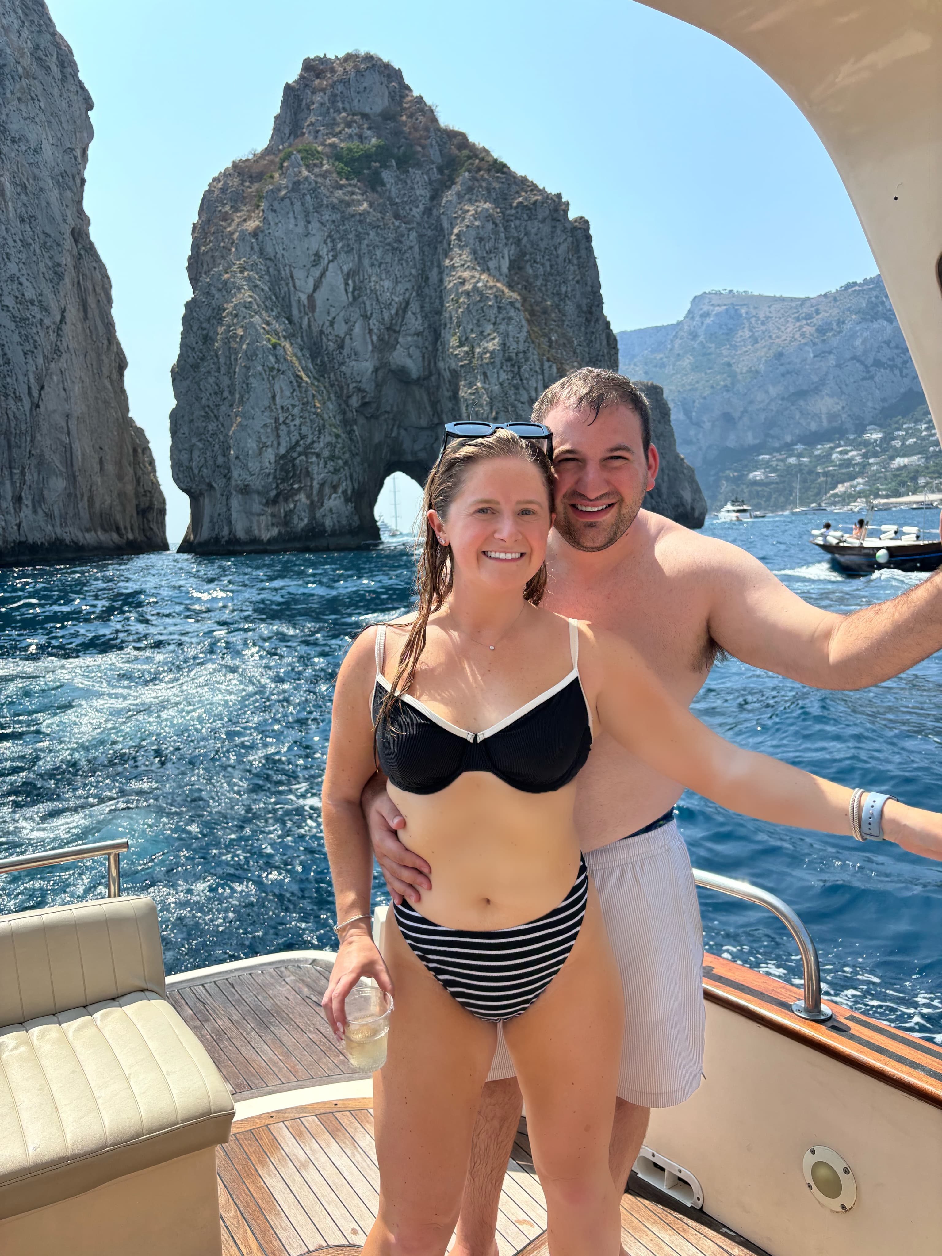 Advisor and husband in swimsuits on a boat with large rock formations behind them on a sunny day