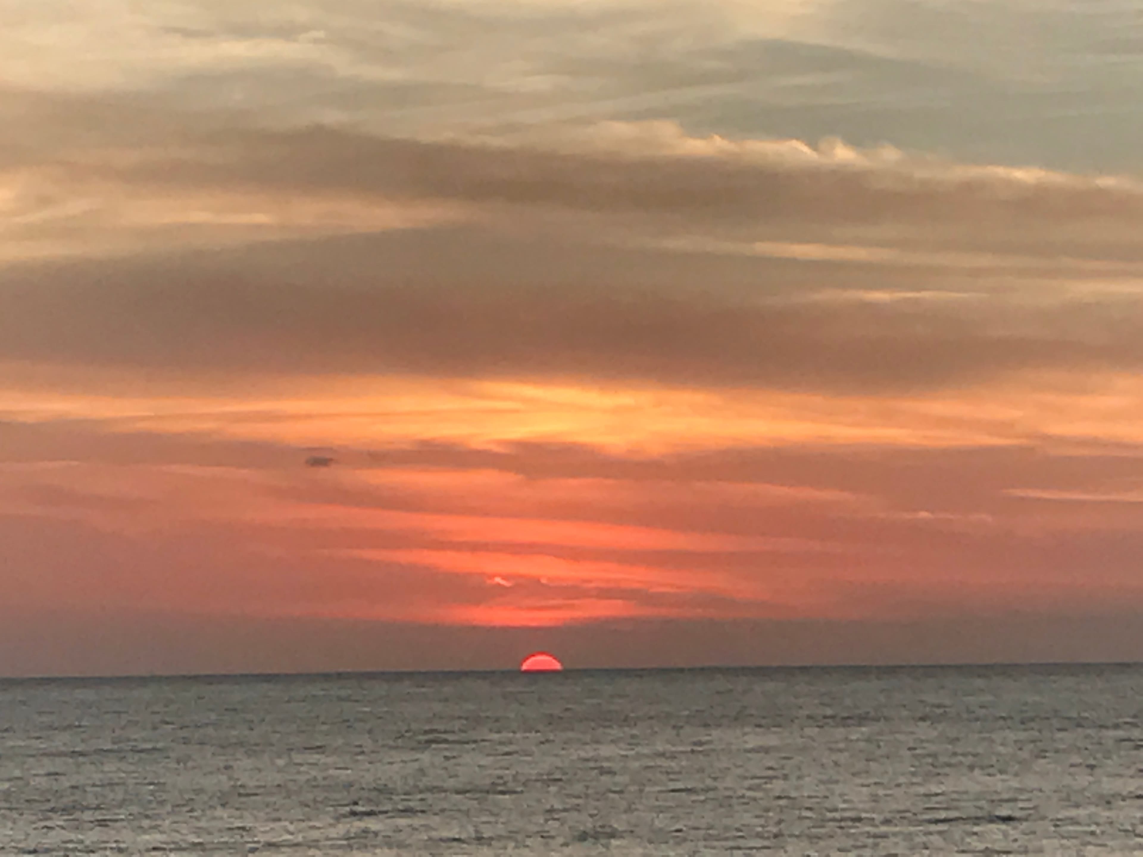 View of the sun halfway below the ocean’s horizon and orange light shining through the clouds above