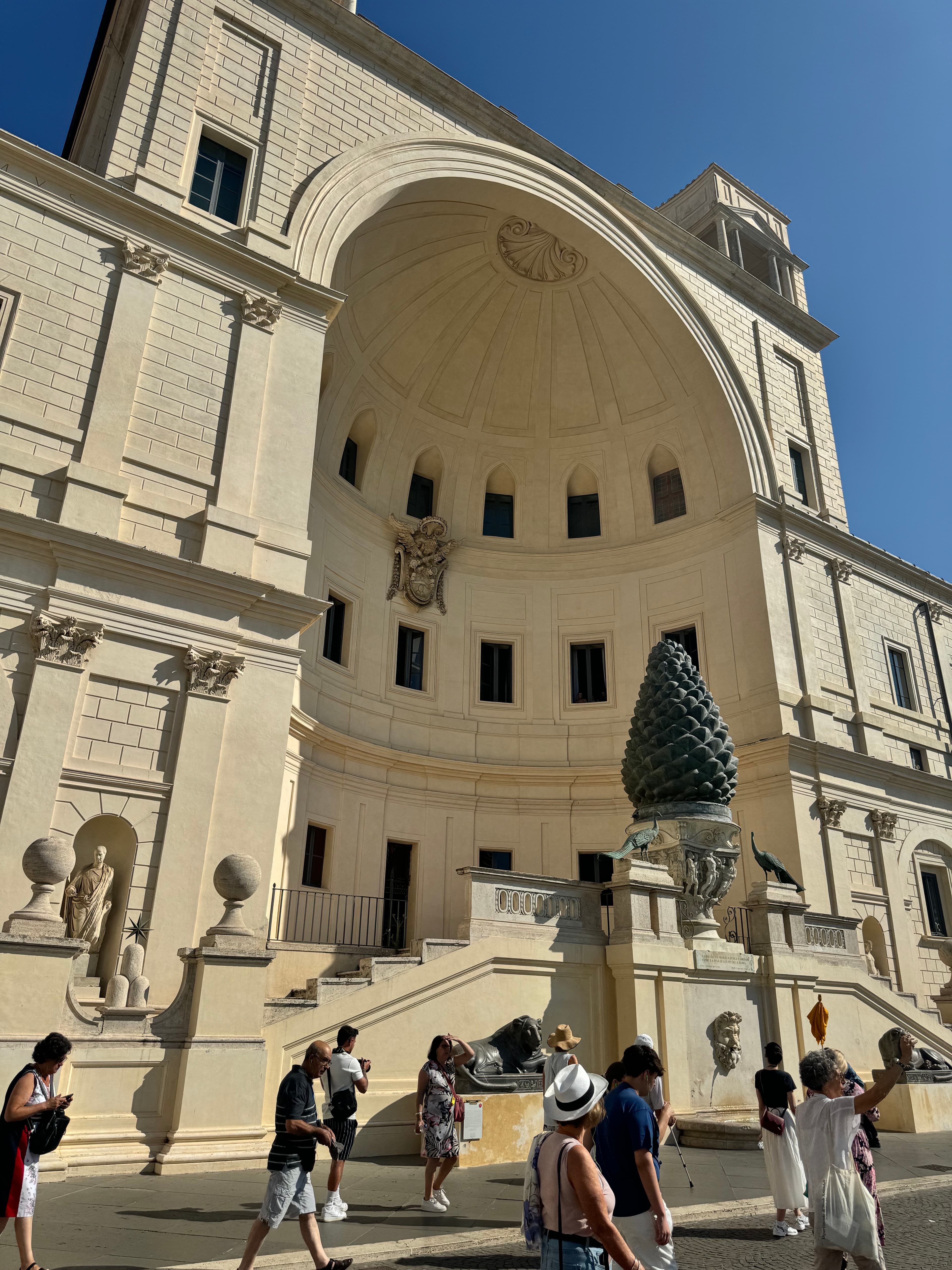 View of a large white building in a city with staircase and sculpture in front and pedestrians walking on the street below