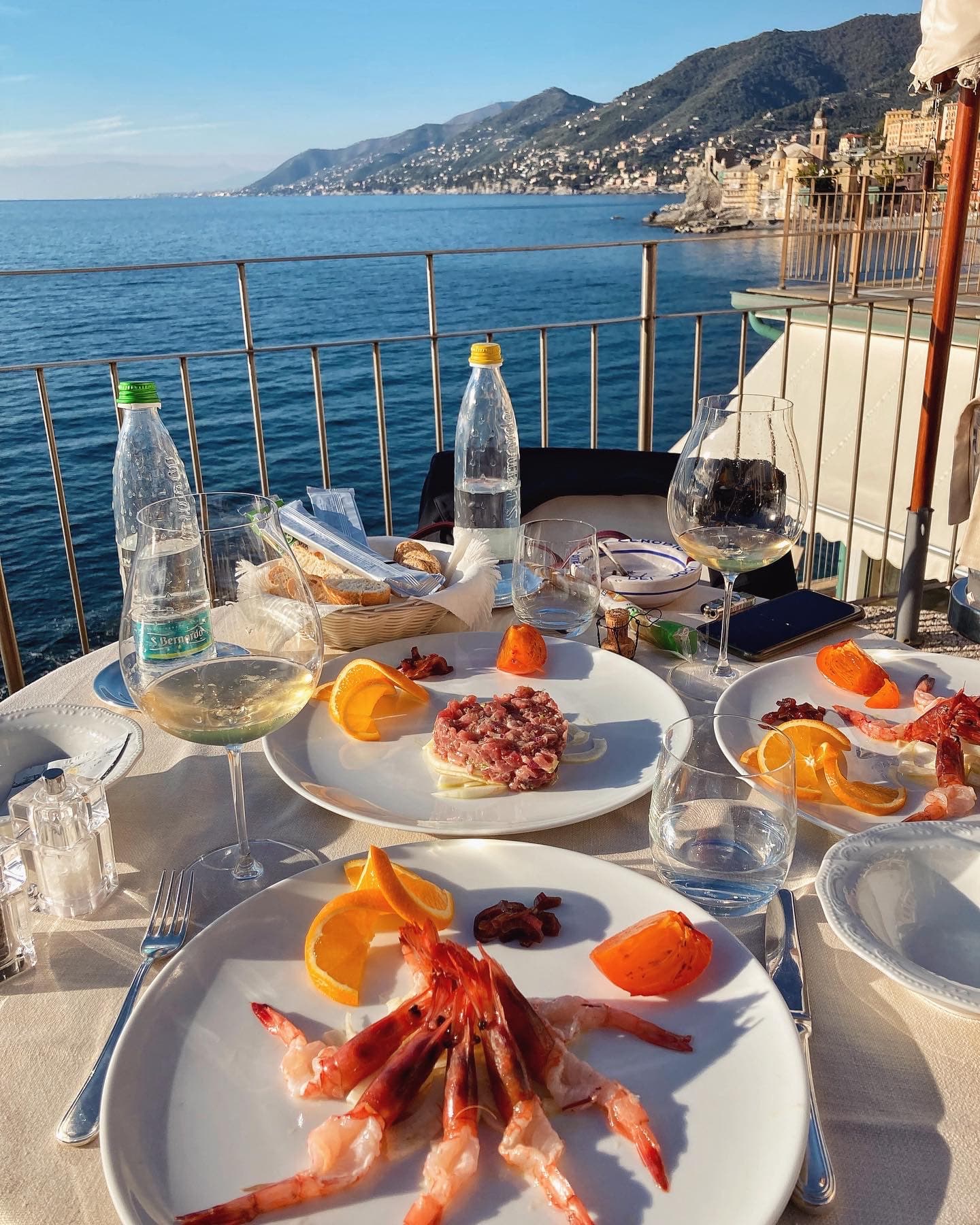 A view of a table filled with plates of seafood, wine glasses and bottled water overlooking a calm ocean and city along the coastline.