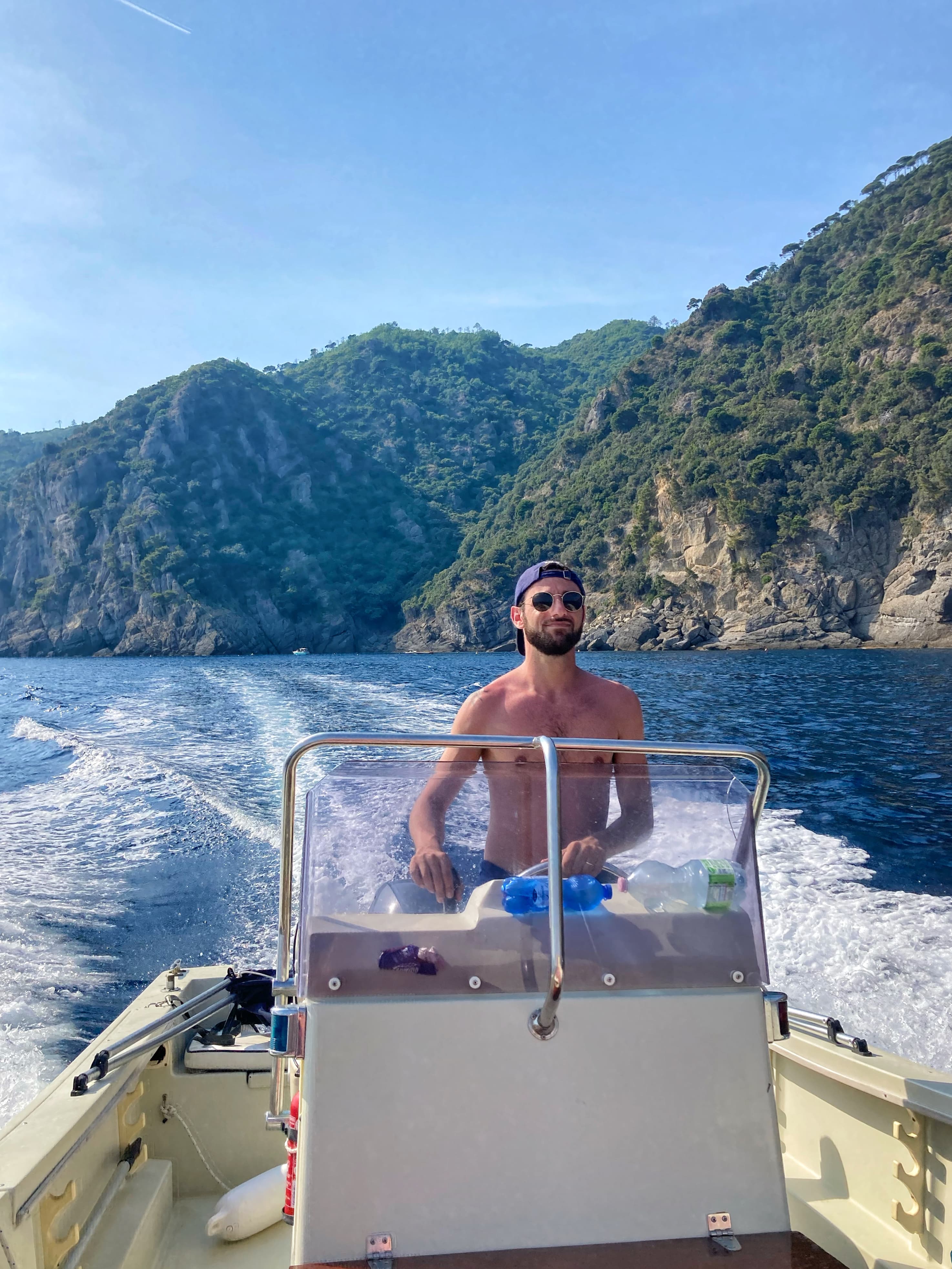 A man driving a boat in the ocean along the mountainous coastline on a sunny day.