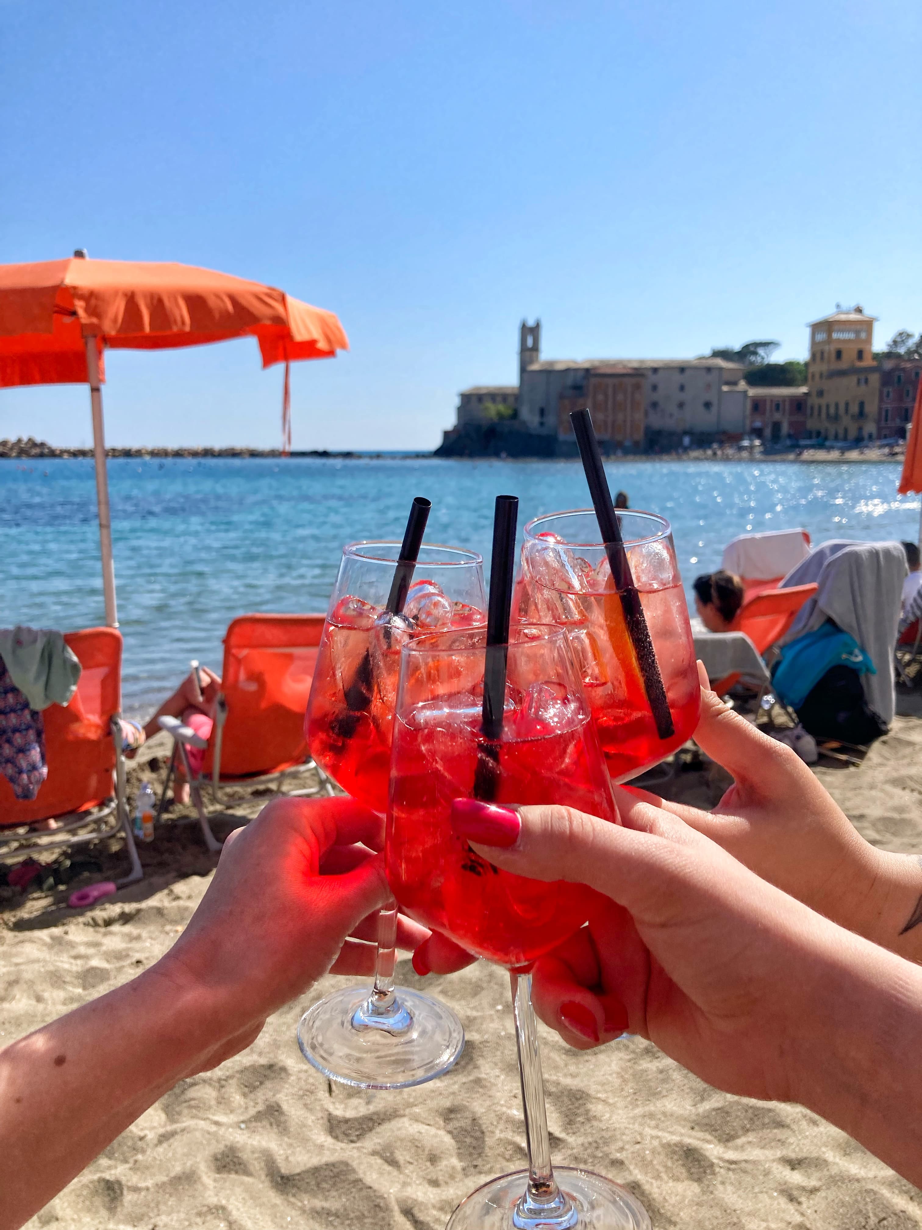 Hands holding red drinks together in a cheers position with a beach scene in the background on a sunny day.