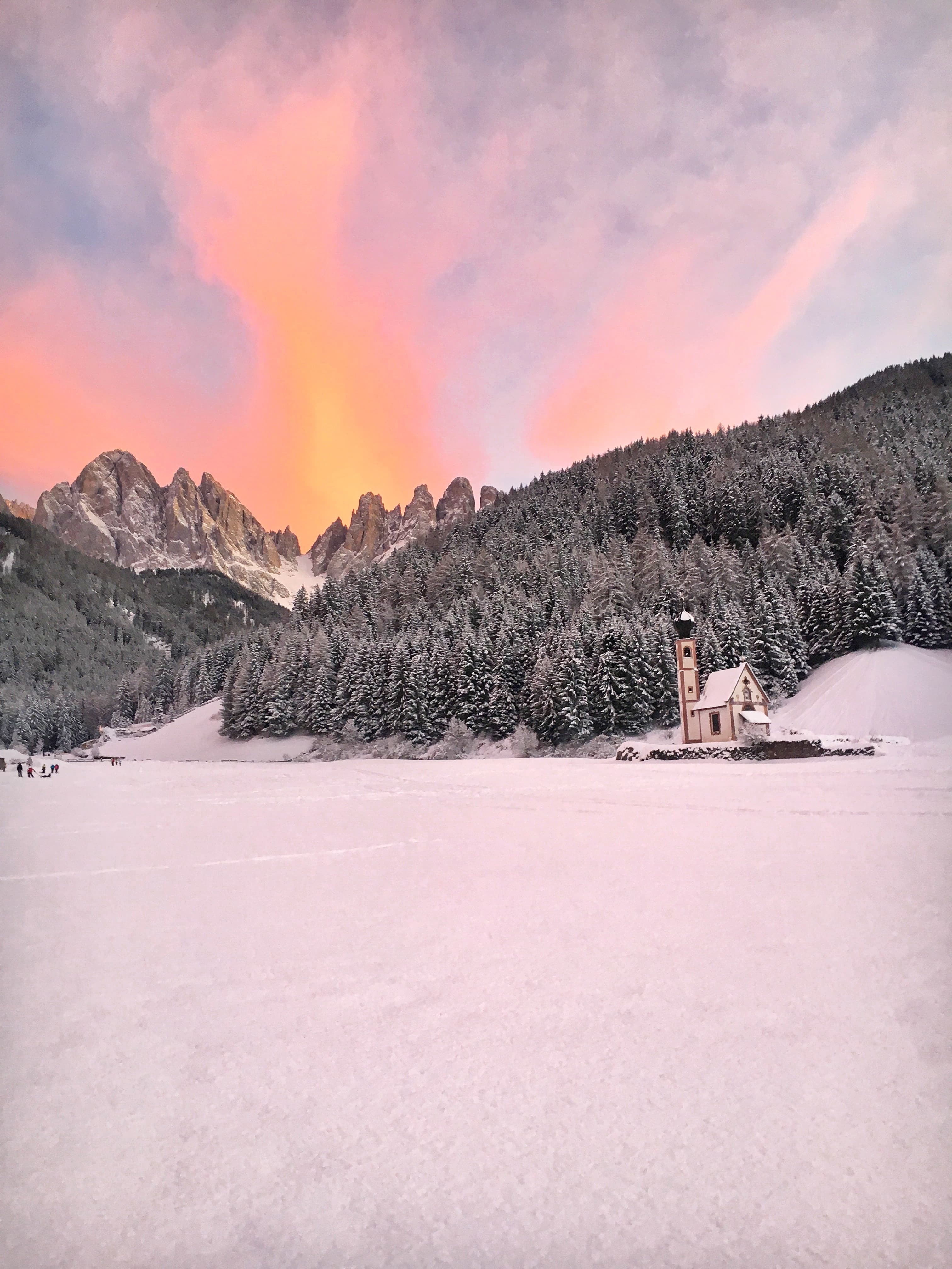 A snow scene with tree covered mountains in the distance and a beautiful orange, pink, white and blue sunrise or sunset in the sky.