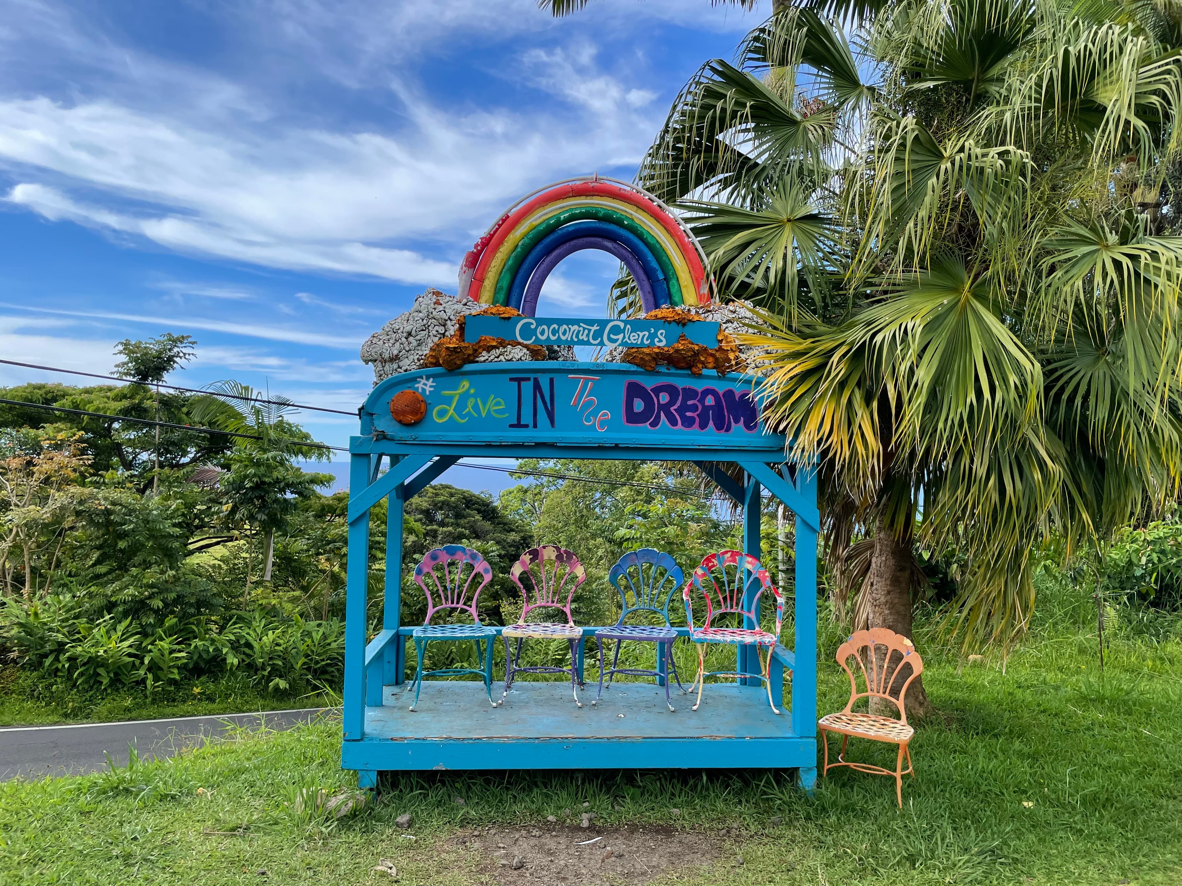 View of a blue structure with a rainbow on top and four chairs near a palm tree on a sunny day