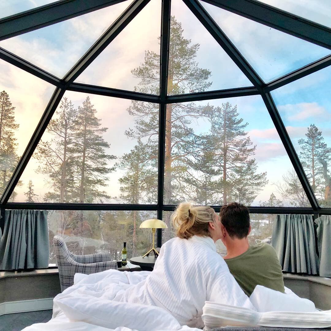 Advisor and partner sitting on a bed inside a glass igloo with tall pine trees visible outdoors