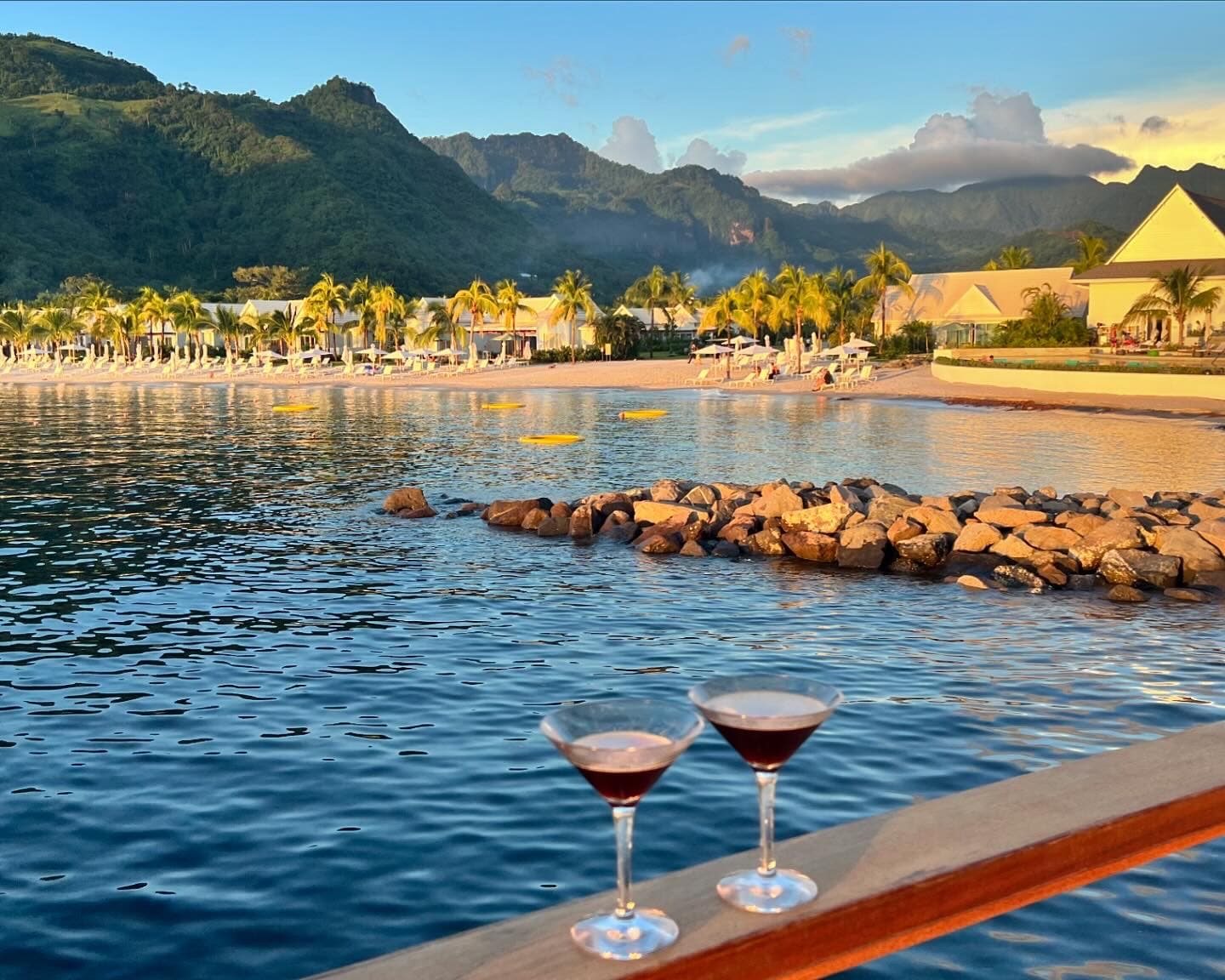 View of two martini glasses sitting on a ledge by a beautiful beach with lush mountains in the distance