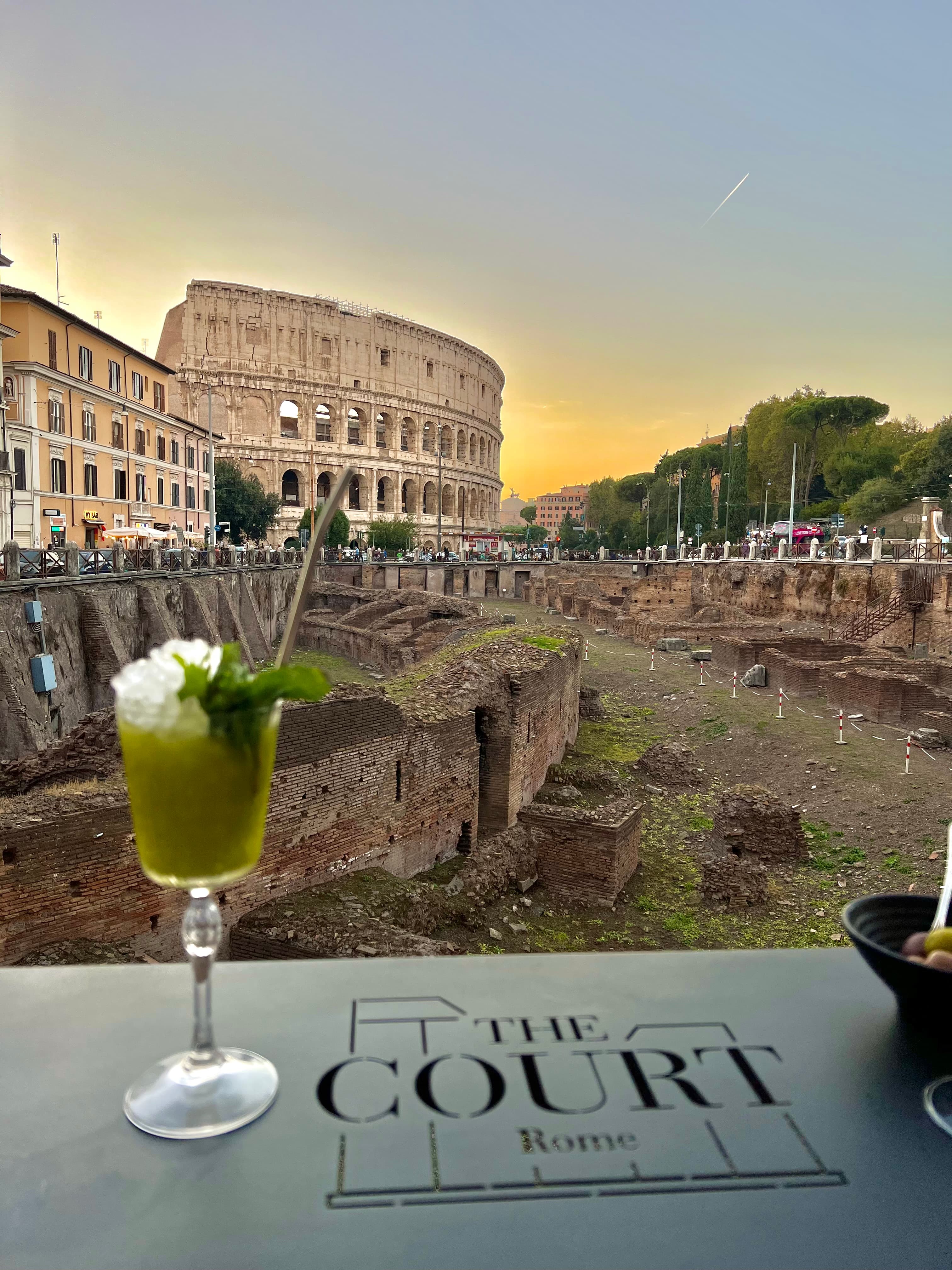 View of a green cocktail on a table at a restaurant with the Colosseum in view at sunset
