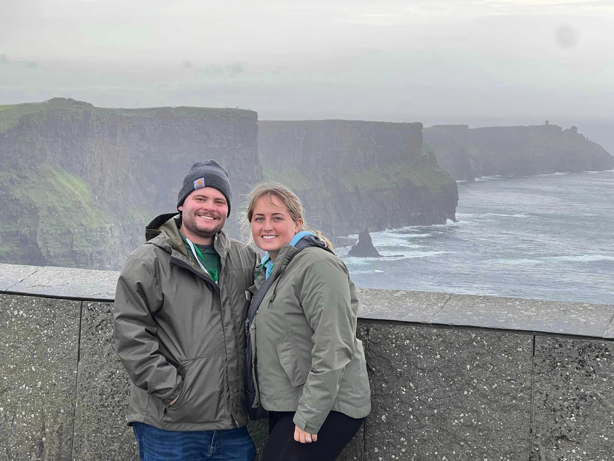 Two people posing for a photo together with cliffs next to the water in the background