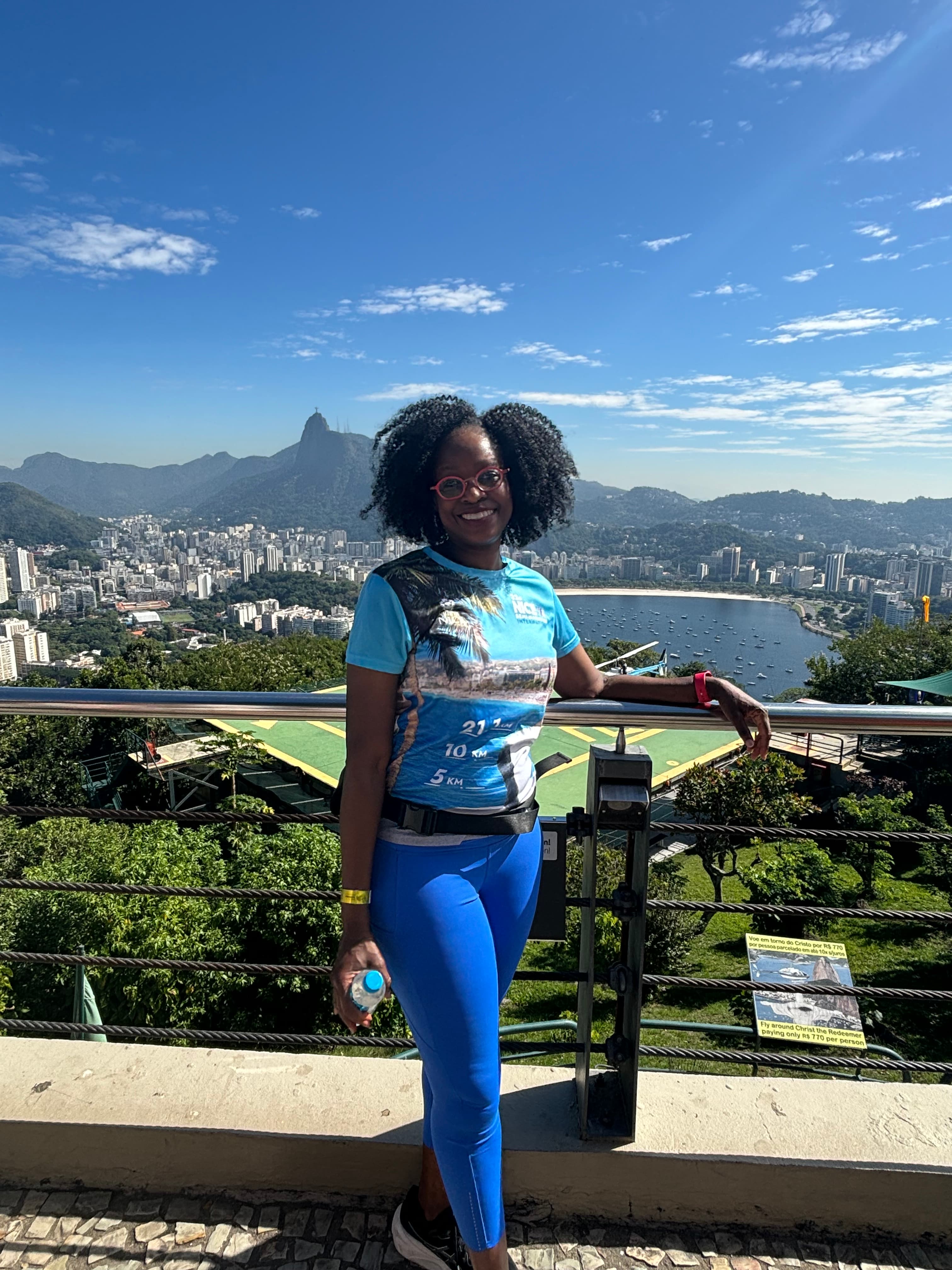 A person standing and smiling for a photograph in front of a fence with an aerial view of a town and mountains in the background