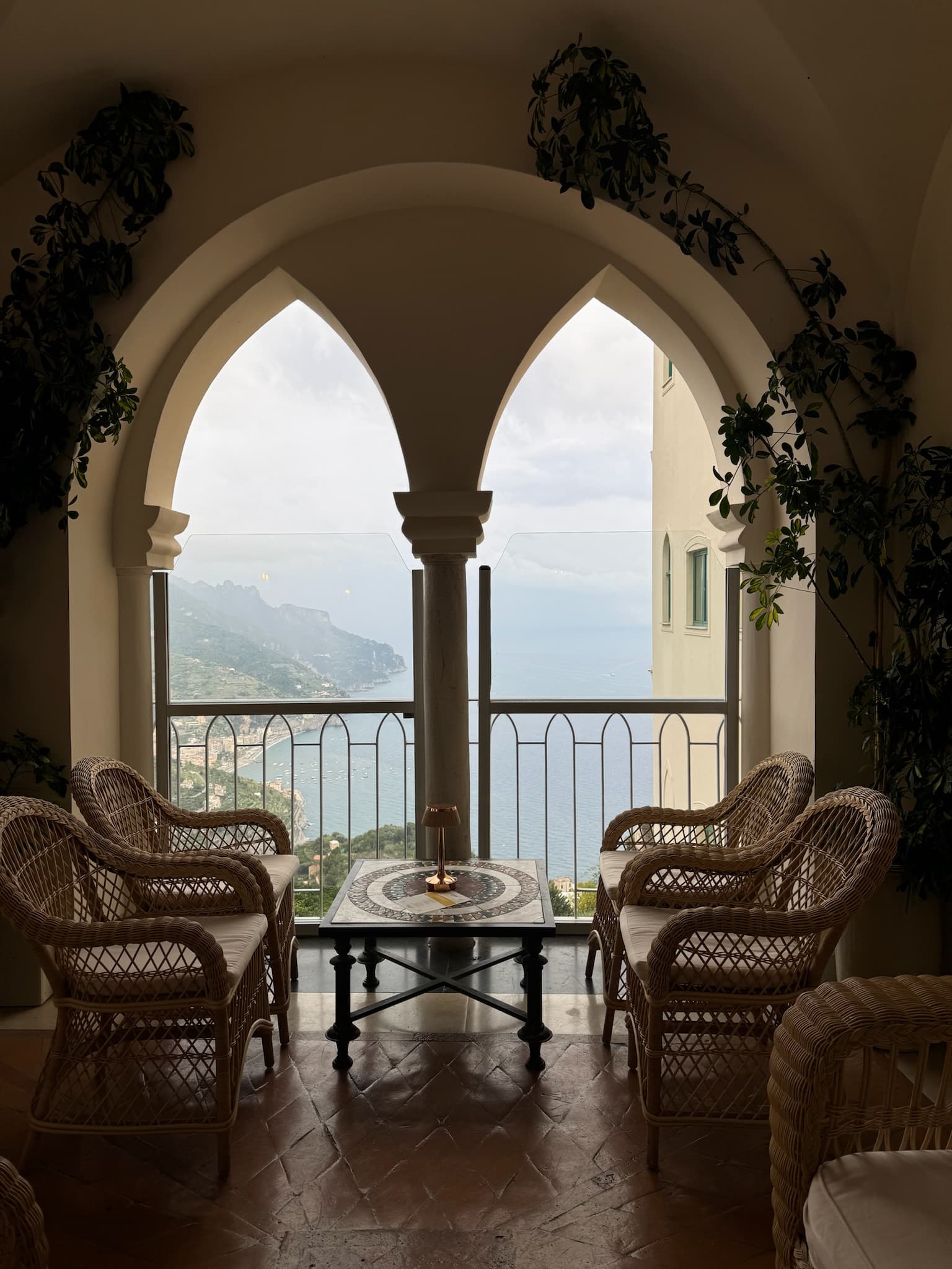 Four wicker chairs and a table set by an open arched window overlooking the coast on a cloudy day