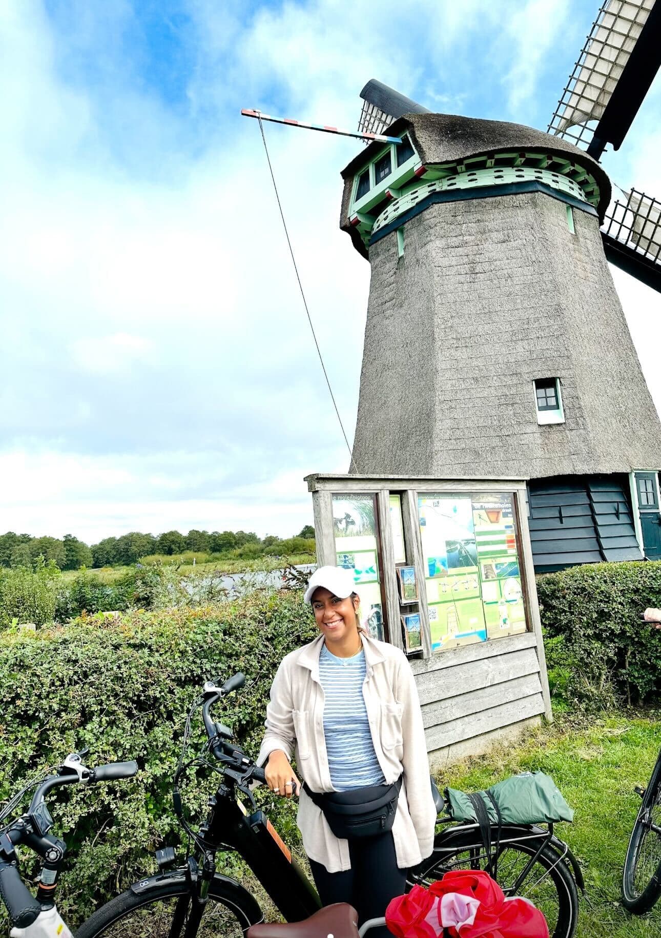 Alexa smiling and posing on a bike with a windmill in the background during the daytime.