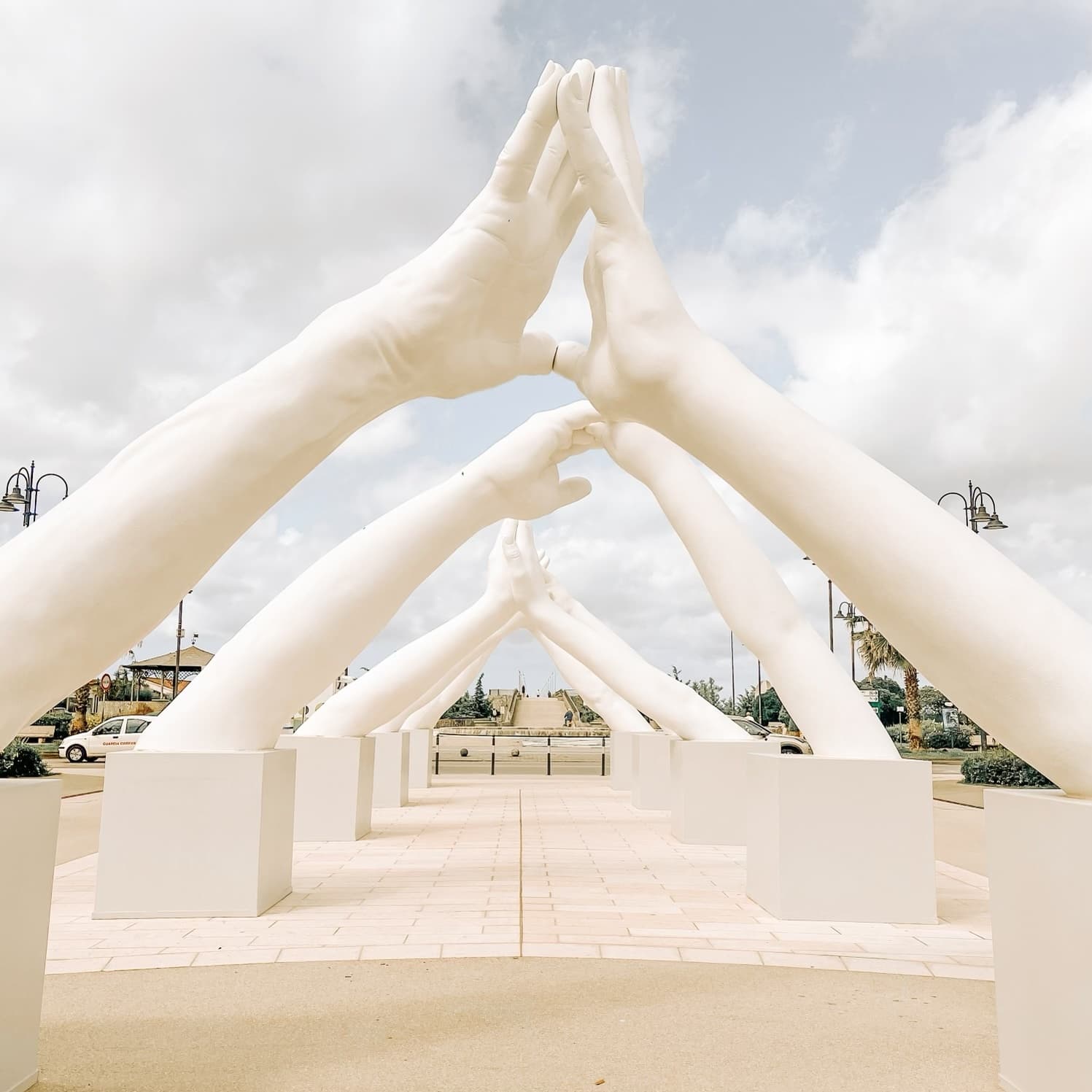 View of large white sculptures of arms touching hands forming a pathway in a sunny city plaza