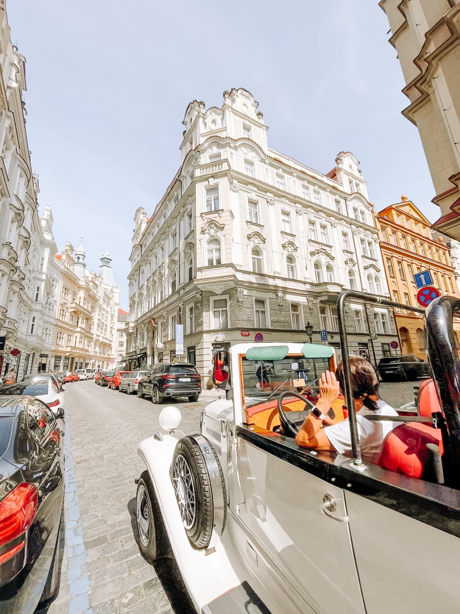 View of an old white car driving on a city street on a sunny day in Prague