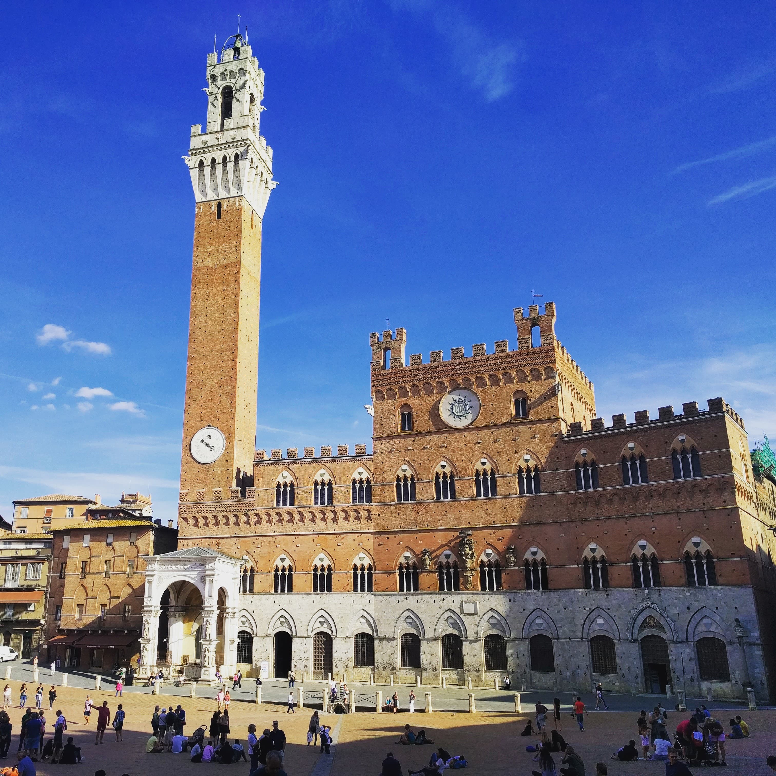 A view of a large historical brick building with a tall tower and tourist standing around the base on a clear blue day.