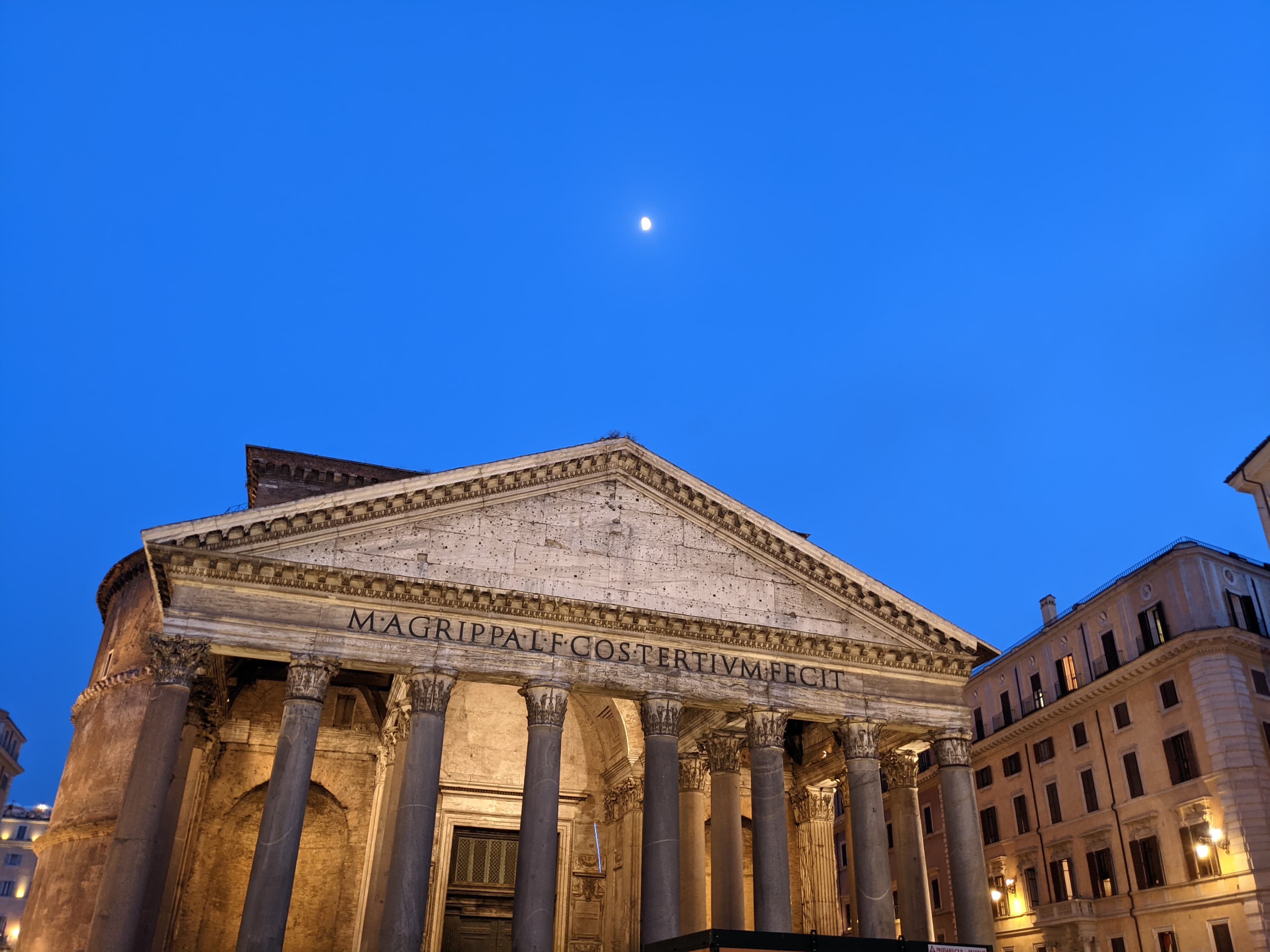 A view of a historical building with white stone pillars at night with the moon above.