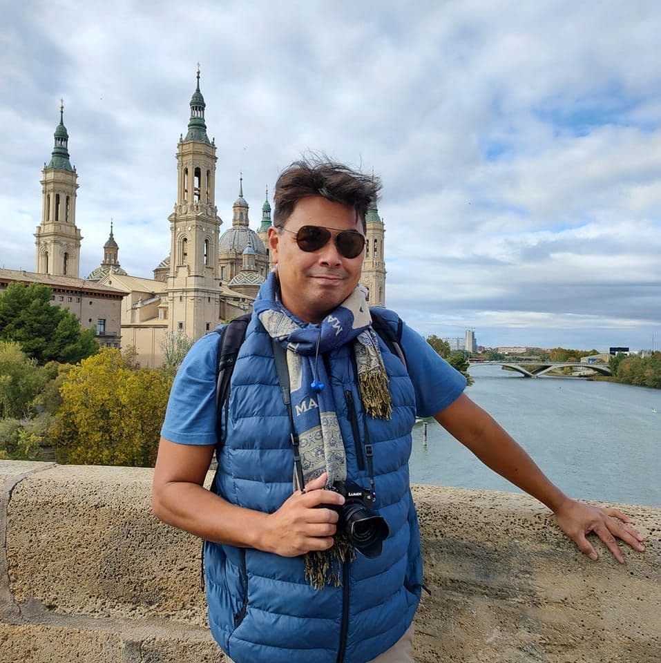 Advisor in a blue vest holding a camera and smiling on a bridge with a beautiful cathedral in the background