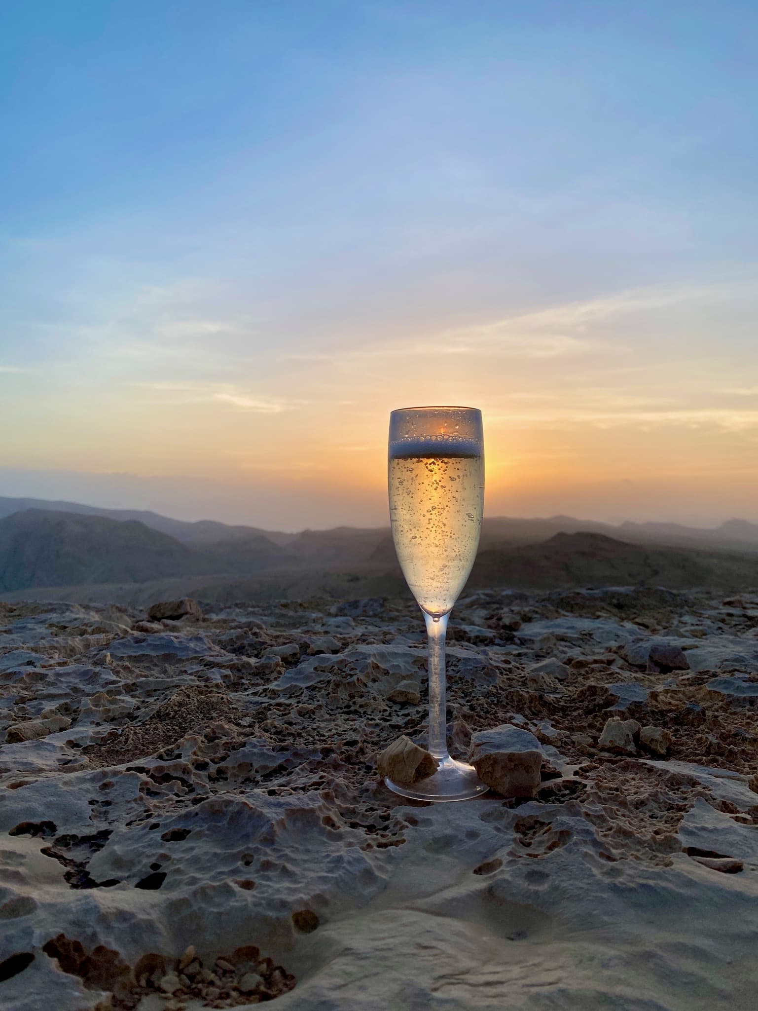 View of a glass of champagne on a rocky surface with a beautiful sunset over the mountains in the distance