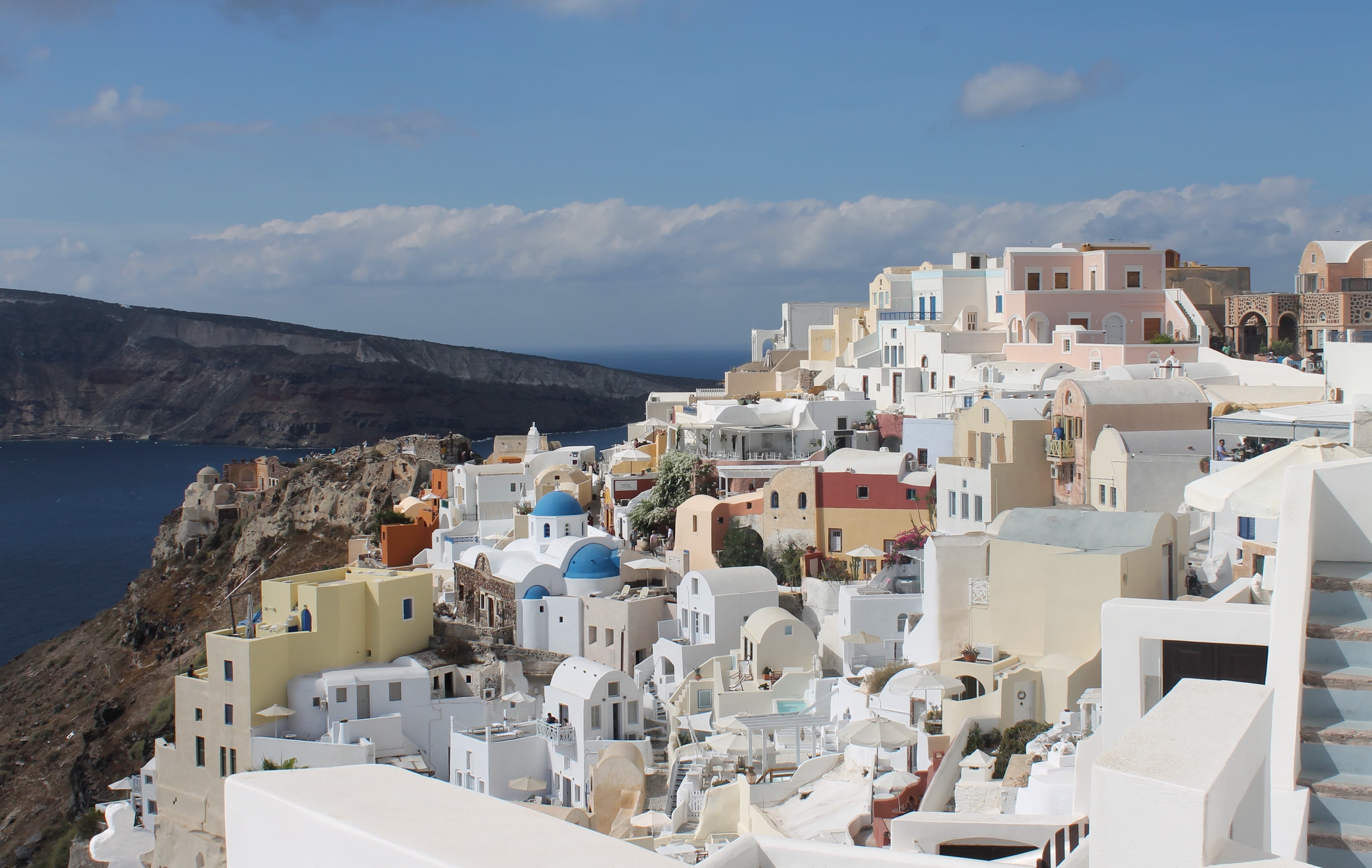 An aerial view of Santorini, Greece with bright white buildings and the famous blue domed rooftops with the water and mountains in the distance on a partly cloudy day.